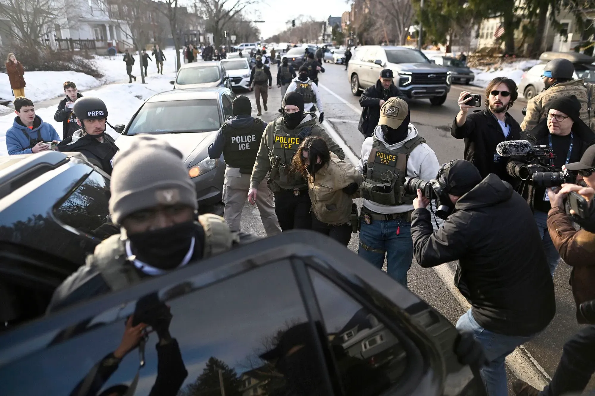 تظهر الصورة مجموعة من رجال الأمن يرتدون زيًا رسميًا، يقومون باعتقال شخص في وسط حشد من الصحفيين والمتفرجين، مما يعكس التوترات حول قضايا الهجرة.