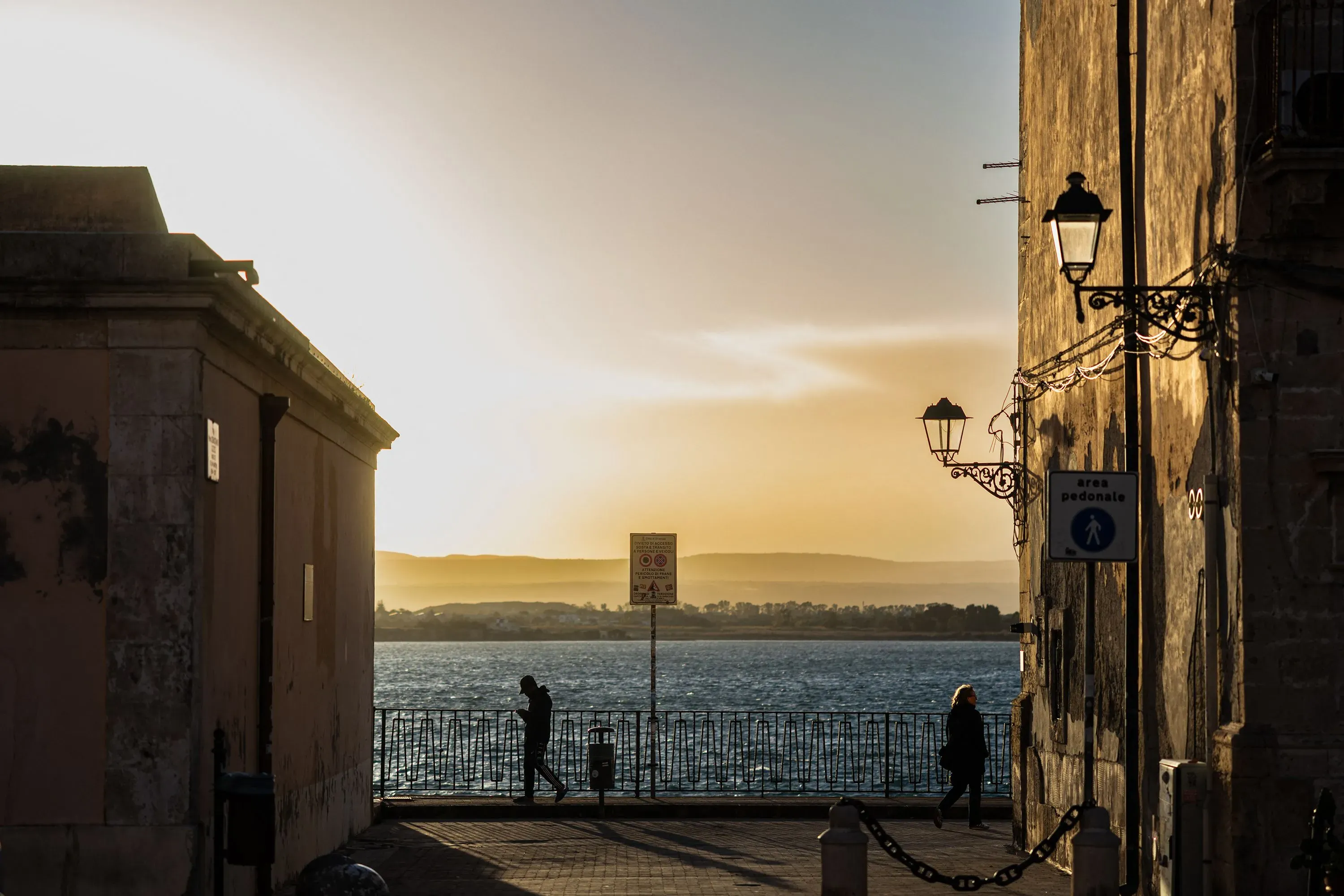 منظر لغروب الشمس على البحر، حيث يسير شخصان على رصيف بجوار مباني تاريخية، مما يعكس جمال الثقافة الإيطالية وتاريخ الهجرة.
