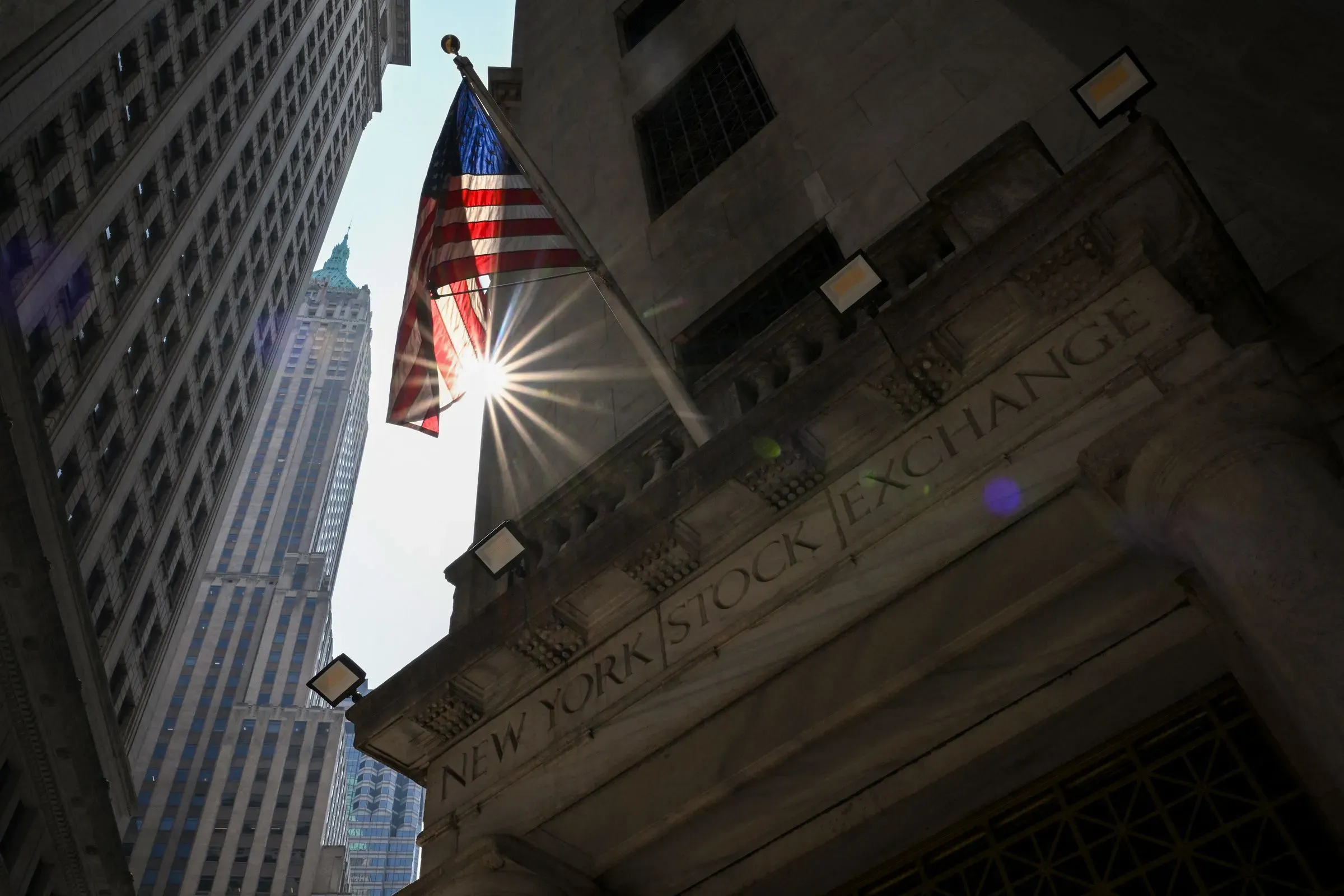 صورة لمبنى بورصة نيويورك مع العلم الأمريكي يرفرف في الأعلى، تظهر أشعة الشمس تتسلل بين المباني العالية، مما يبرز أهمية الأسواق المالية.