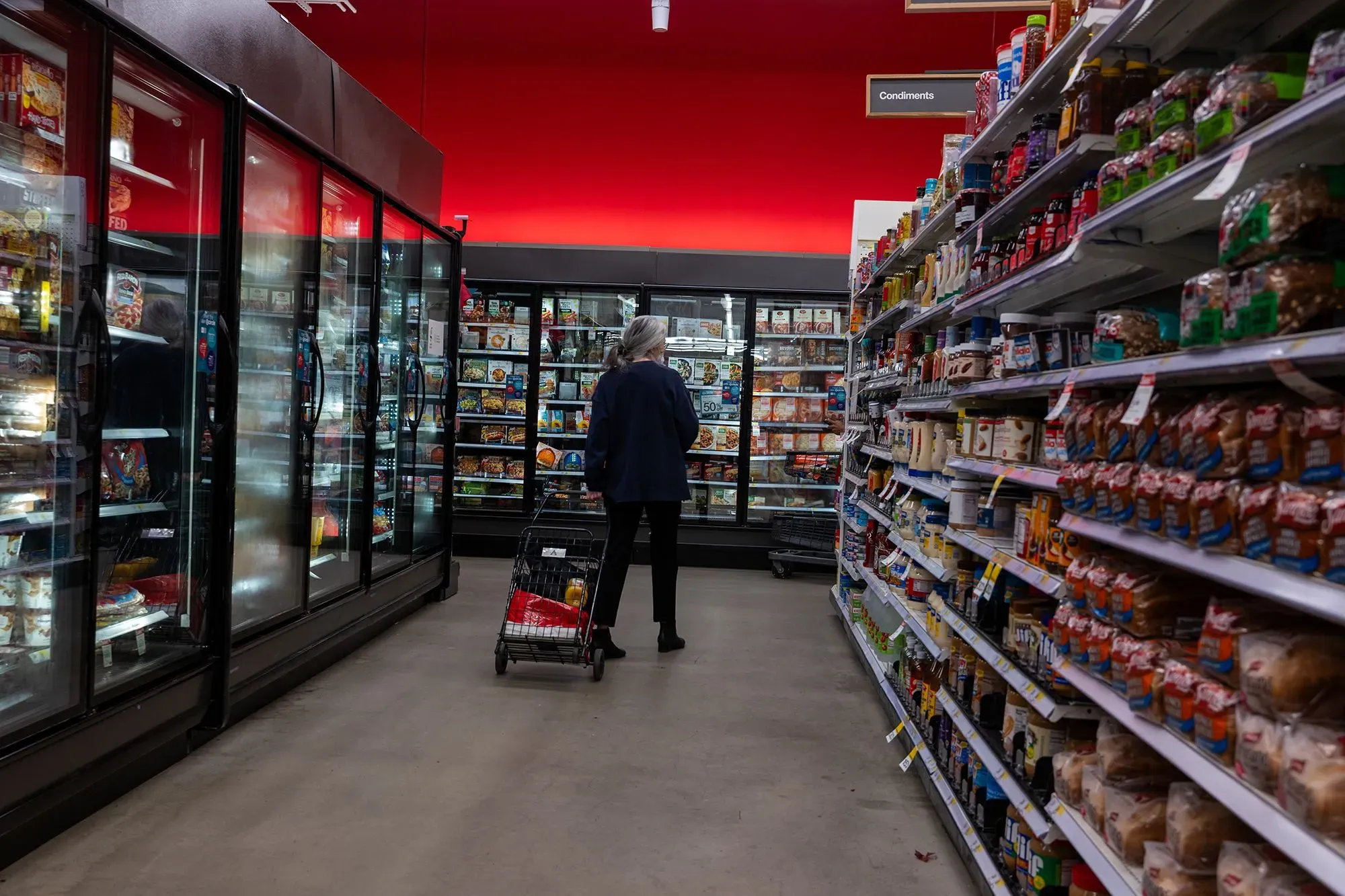 امرأة تتسوق في ممر سوبرماركت، محاطة بأطعمة معالجة ومجمدة، مع خلفية حمراء تُبرز المنتجات. تعكس الصورة التوجه نحو الأطعمة الطبيعية.