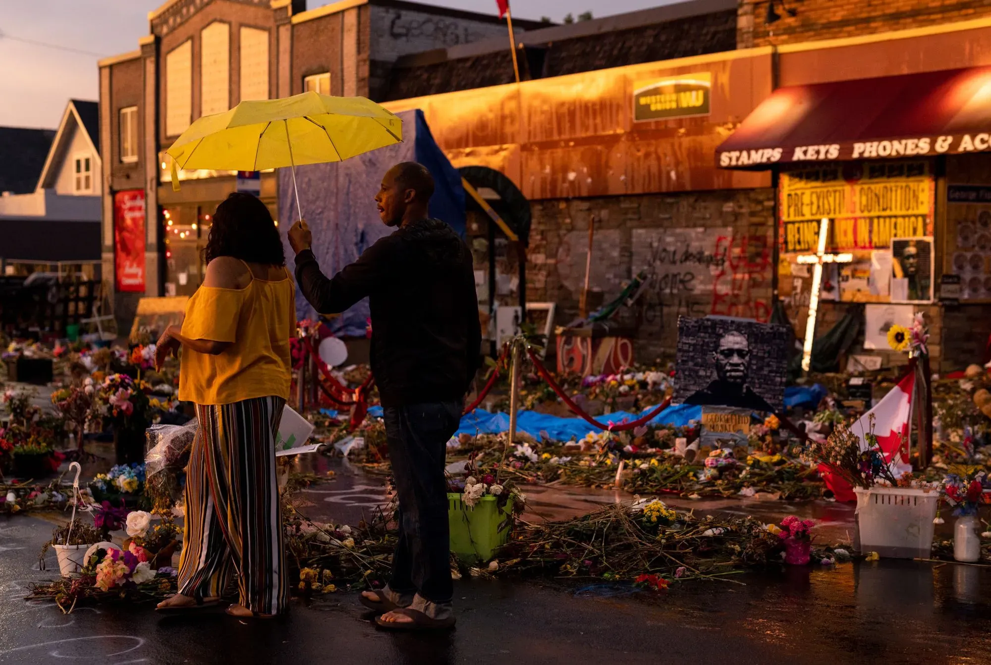 موقع تذكاري في مينيابوليس يضم زهورًا وصورًا تكريمًا لضحايا العنف، مع شخصين يحملان مظلة صفراء في الخلفية.