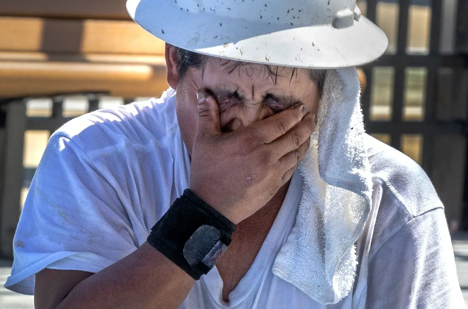 عامل بناء يضع يده على وجهه بسبب الحرارة الشديدة، مرتديًا قبعة وقميصًا أبيض، مع منشفة على كتفه، تعبيره يعكس المعاناة من الرطوبة والحرارة.