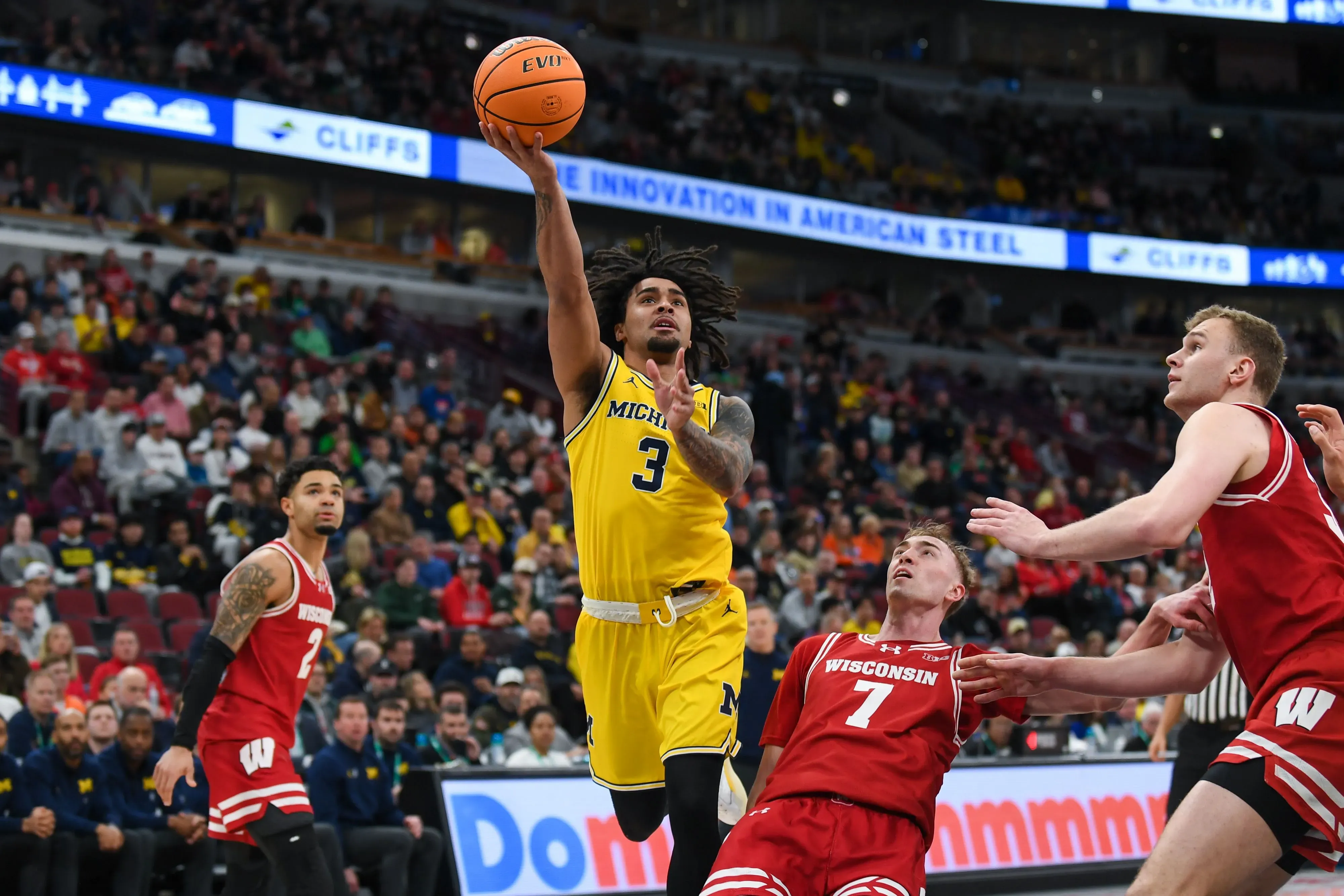 لاعب كرة سلة من فريق ميشيغان يقوم بقفزة رائعة لتسديد الكرة، بينما يتنافس مع لاعبين من فريق ويسكونسن في مباراة حماسية.