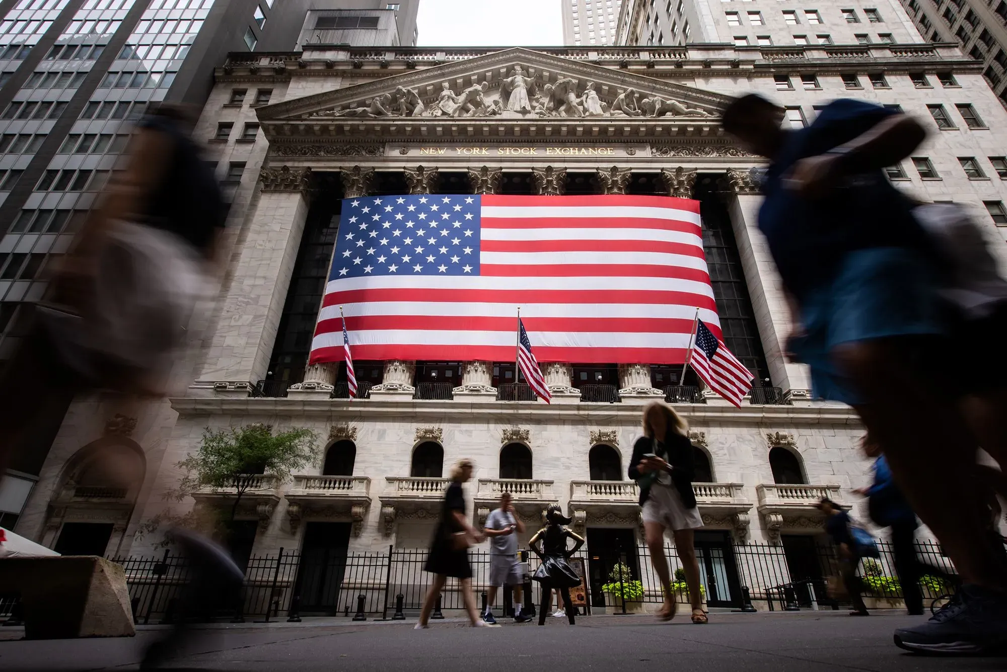 واجهة بورصة نيويورك مزينة بعلم الولايات المتحدة، مع مشاة يتجولون في الشارع، تعكس النشاط الاقتصادي والاهتمام بالأسواق المالية.
