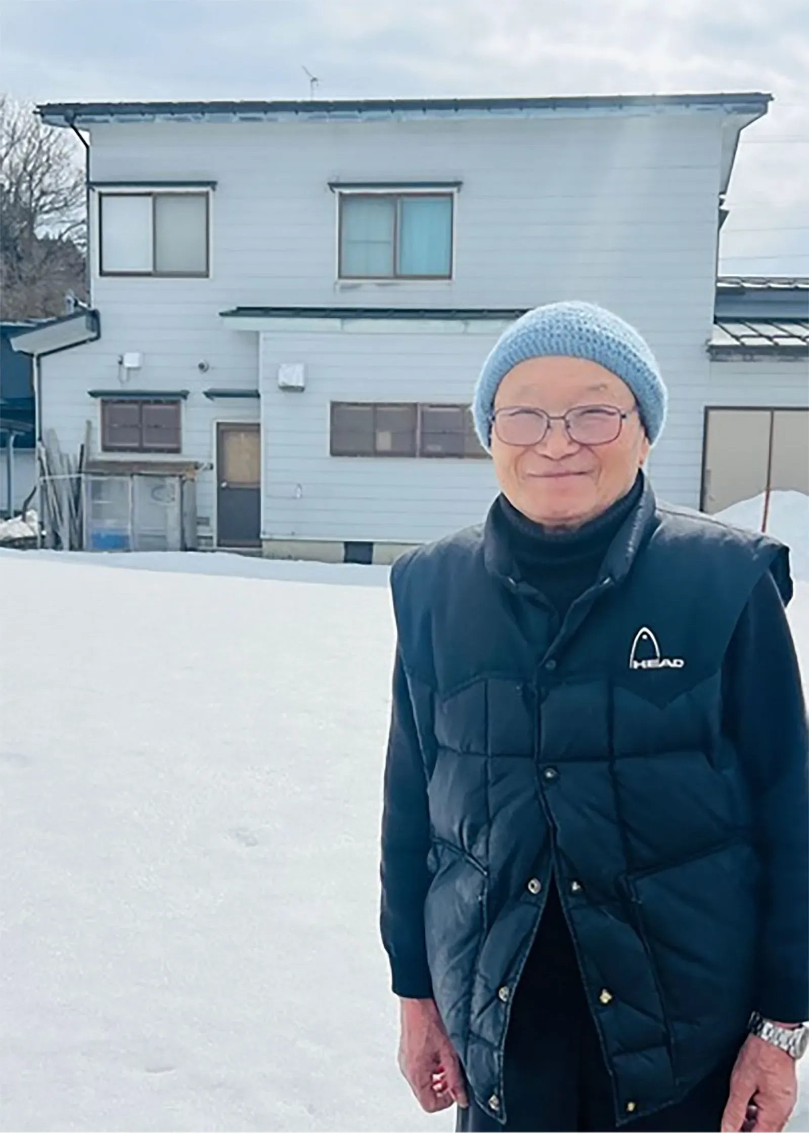 امرأة مسنّة ترتدي سترة داكنة وقبعة صوفية، تقف أمام منزلها المغطى بالثلوج في شمال اليابان، تعكس تحديات الشتاء القاسي.