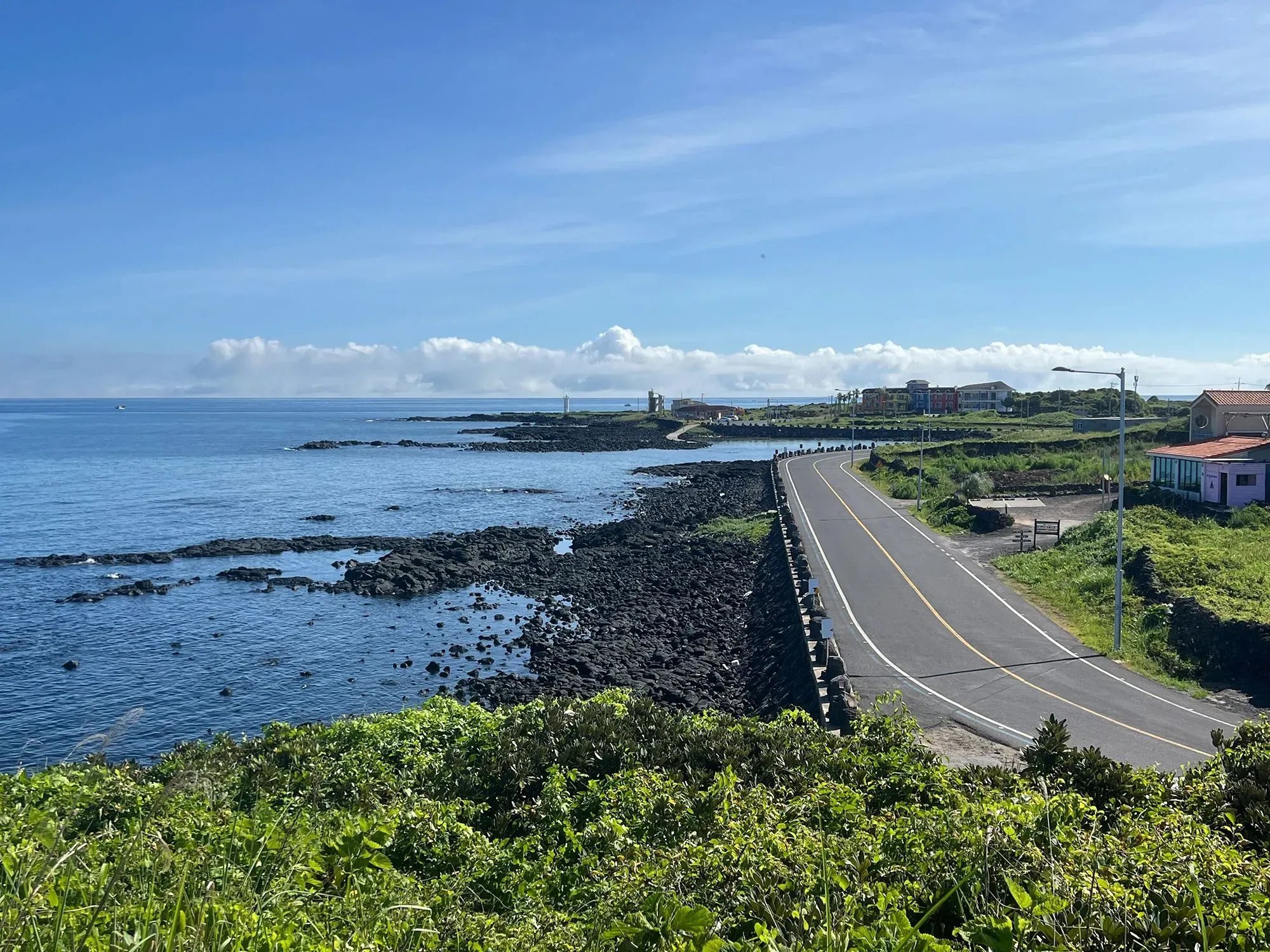 منظر ساحلي لجزيرة جيجو يظهر الطريق المحاذي للشاطئ والمياه الزرقاء المتلألئة، مع خلفية من المباني والمناظر الطبيعية الخضراء.