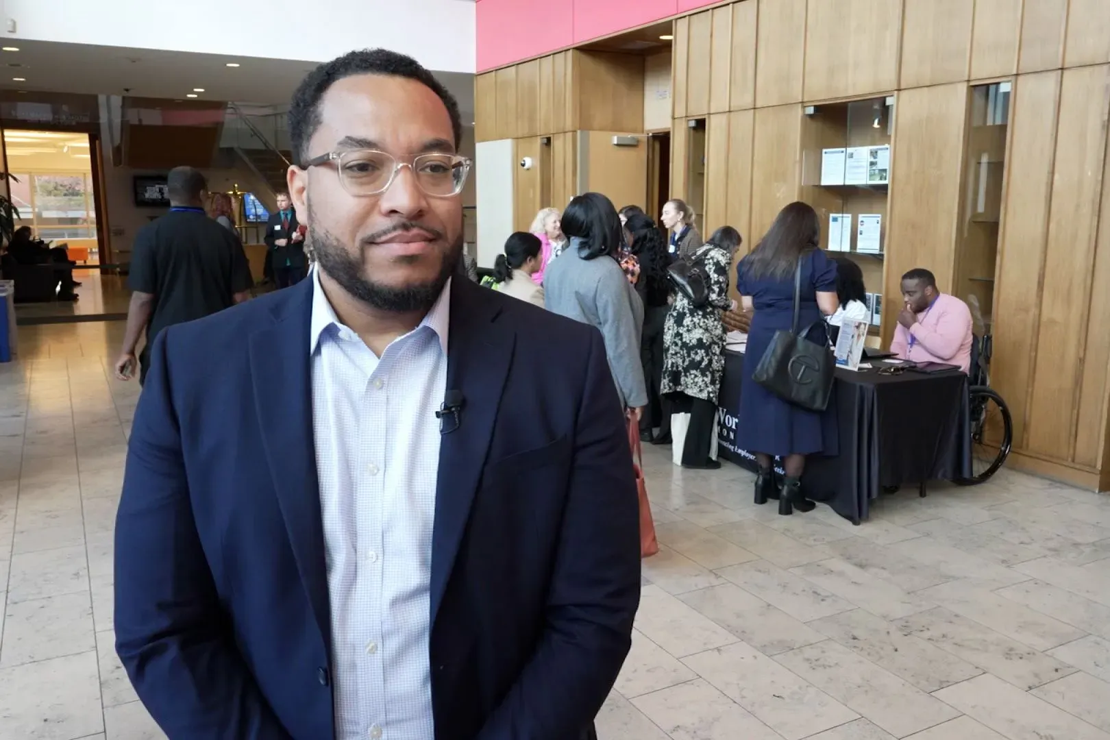 رجل يرتدي بدلة داكنة يقف في مقدمة صالة عرض مع مجموعة من الأشخاص في الخلفية، حيث يشاركون في معرض توظيف.