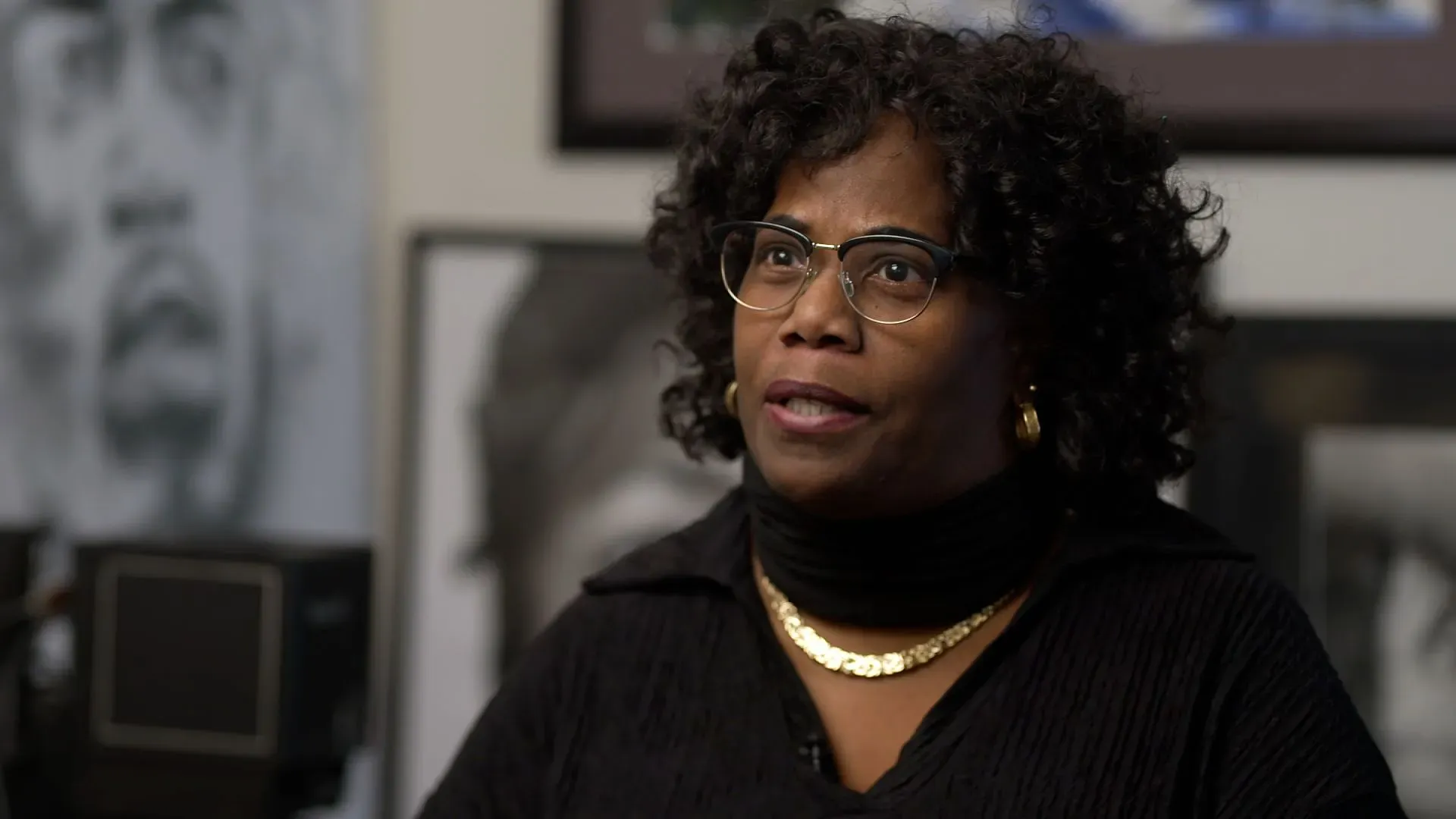 امرأة ذات شعر مجعد ترتدي نظارات وتبتسم، تتحدث في استوديو مليء باللوحات الفنية. تعكس تعبيراتها التحديات التي تواجهها كموظفة حكومية في ظل الإغلاق.