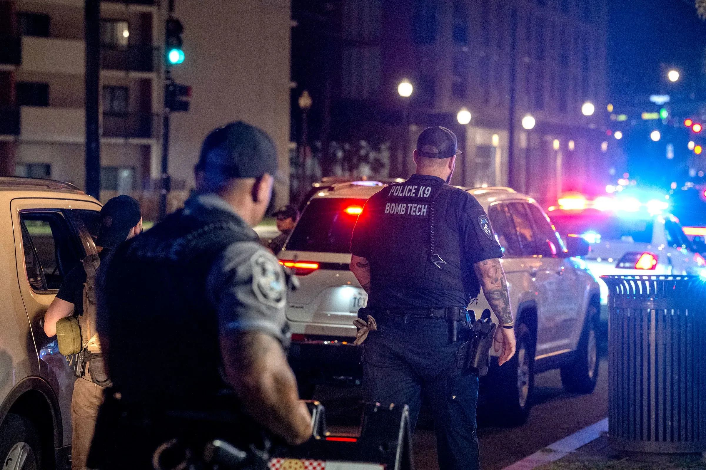 تظهر الصورة ضباط شرطة في العاصمة واشنطن خلال عملية تفتيش ليلي، مع وجود سيارات شرطة مضاءة في الخلفية، مما يعكس تأثير السياسات الأمنية.