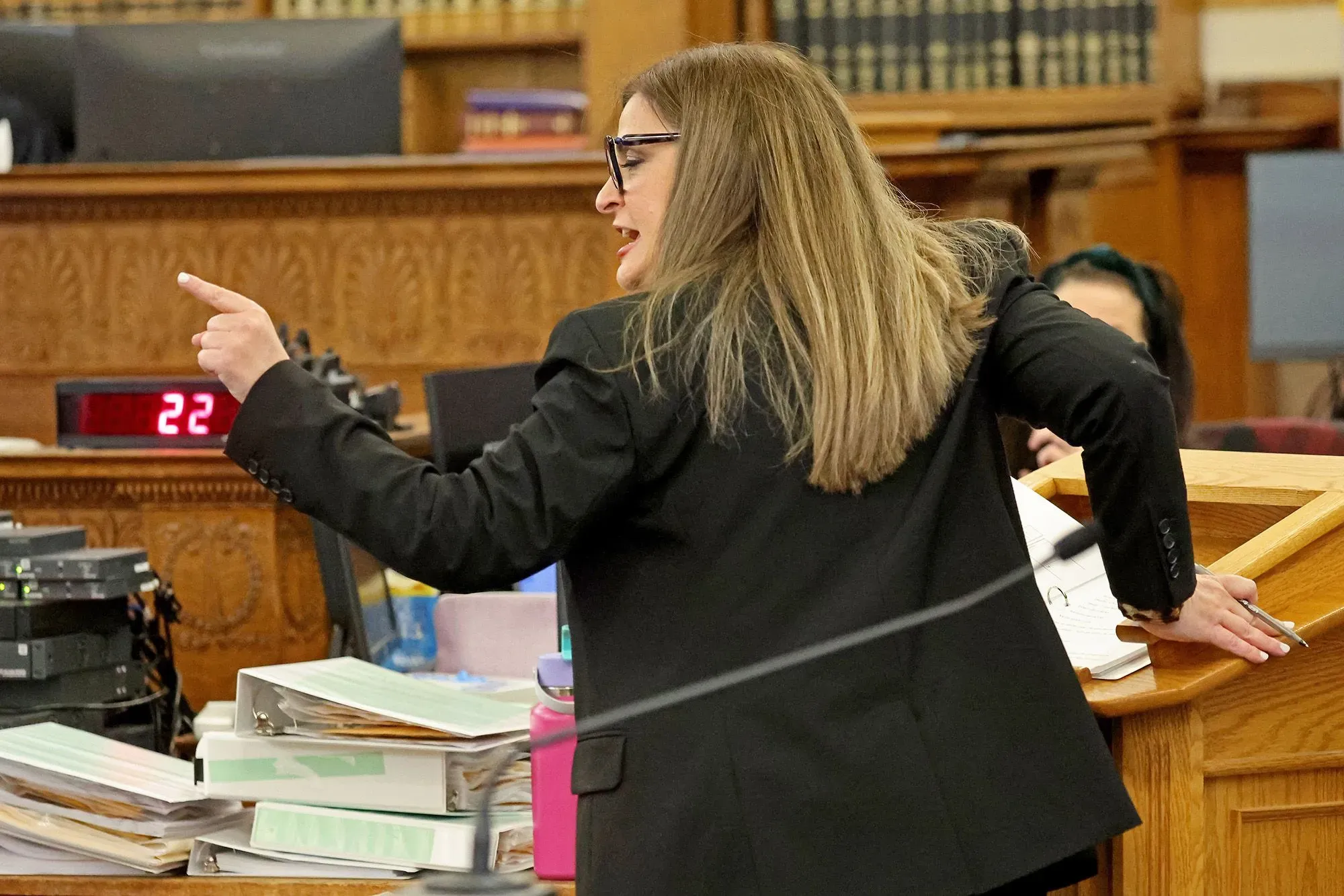 محامية تتحدث بحماس خلال المحاكمة، تشير بيدها نحو هيئة المحلفين، مع وجود مستندات وأجهزة في الخلفية.