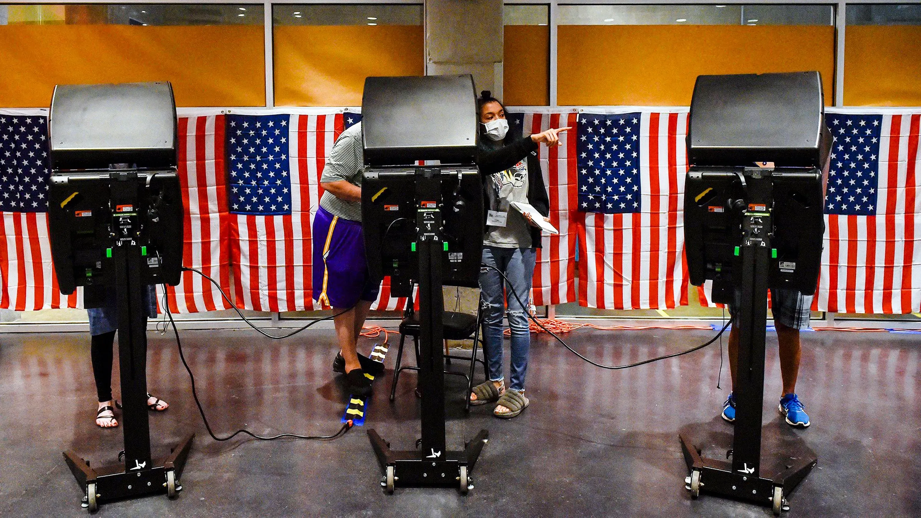 معدات التصويت في مركز اقتراع أمريكي، مع أشخاص يستخدمون الآلات، وخلفهم علم الولايات المتحدة، مما يعكس أهمية الانتخابات.