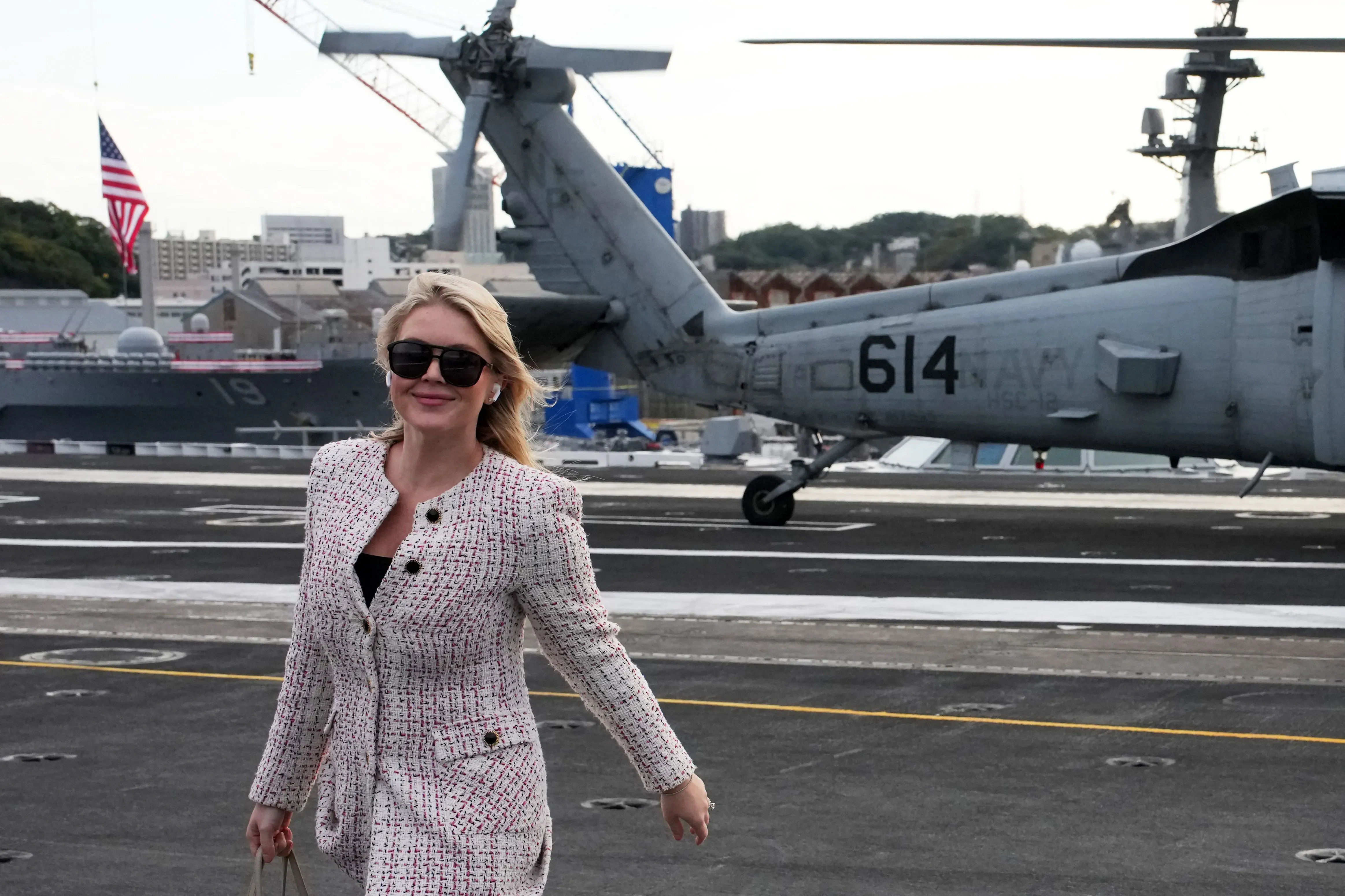 امرأة ترتدي بدلة أنيقة وتبتسم بينما تسير على متن حاملة طائرات، مع طائرات هليكوبتر وسفن في الخلفية، تعكس التنوع الثقافي في كوريا الجنوبية.