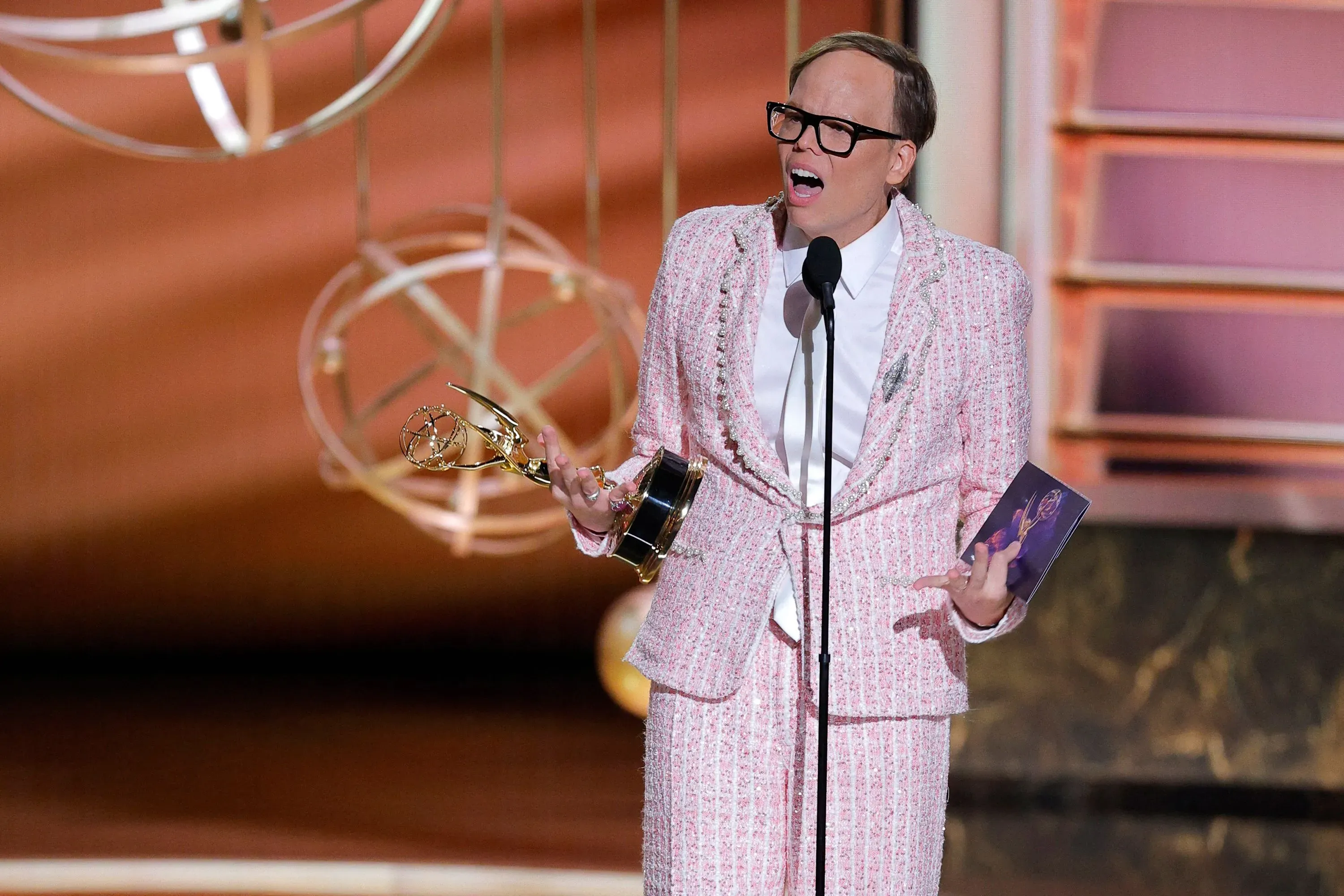 الفائز بجائزة إيمي، جيف هيلر، يتحدث على المسرح مرتديًا بدلة وردية، ممسكًا بجائزته، مع تعبيرات وجه تعكس الفرح والدهشة.