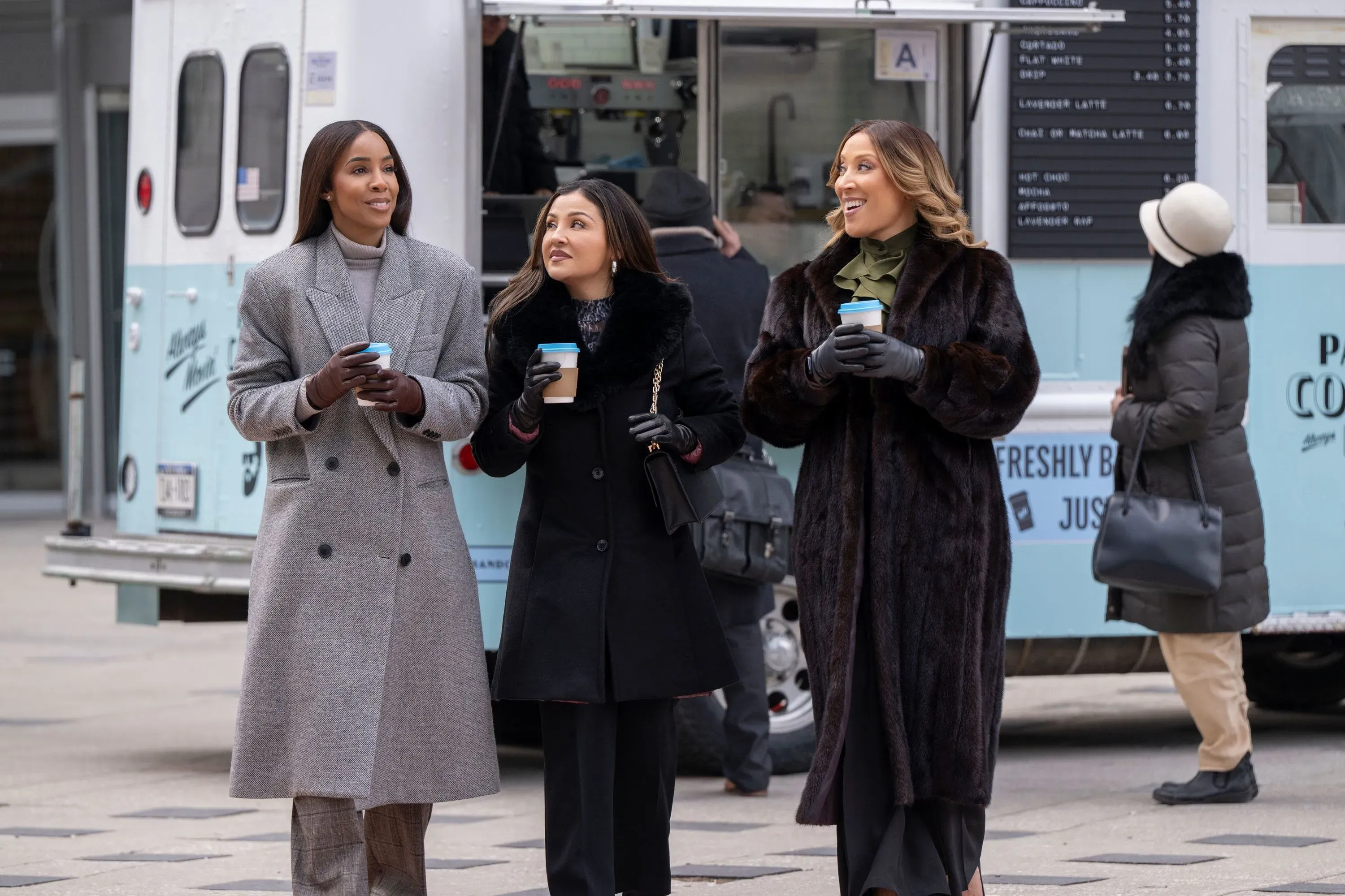 ثلاث نساء يتجولن في الشارع، يحملن أكواب مشروبات ساخنة، بينما يظهر شاحنة طعام في الخلفية، تعكس أجواء الفيلم الرومانسي "أهداف العلاقة".