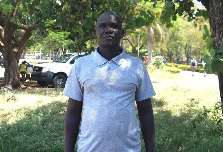 شخص يقف في منطقة خضراء، يرتدي قميصًا رماديًا، مع أشجار في الخلفية، مما يعكس التغيرات الثقافية في مجتمعات الصيد.