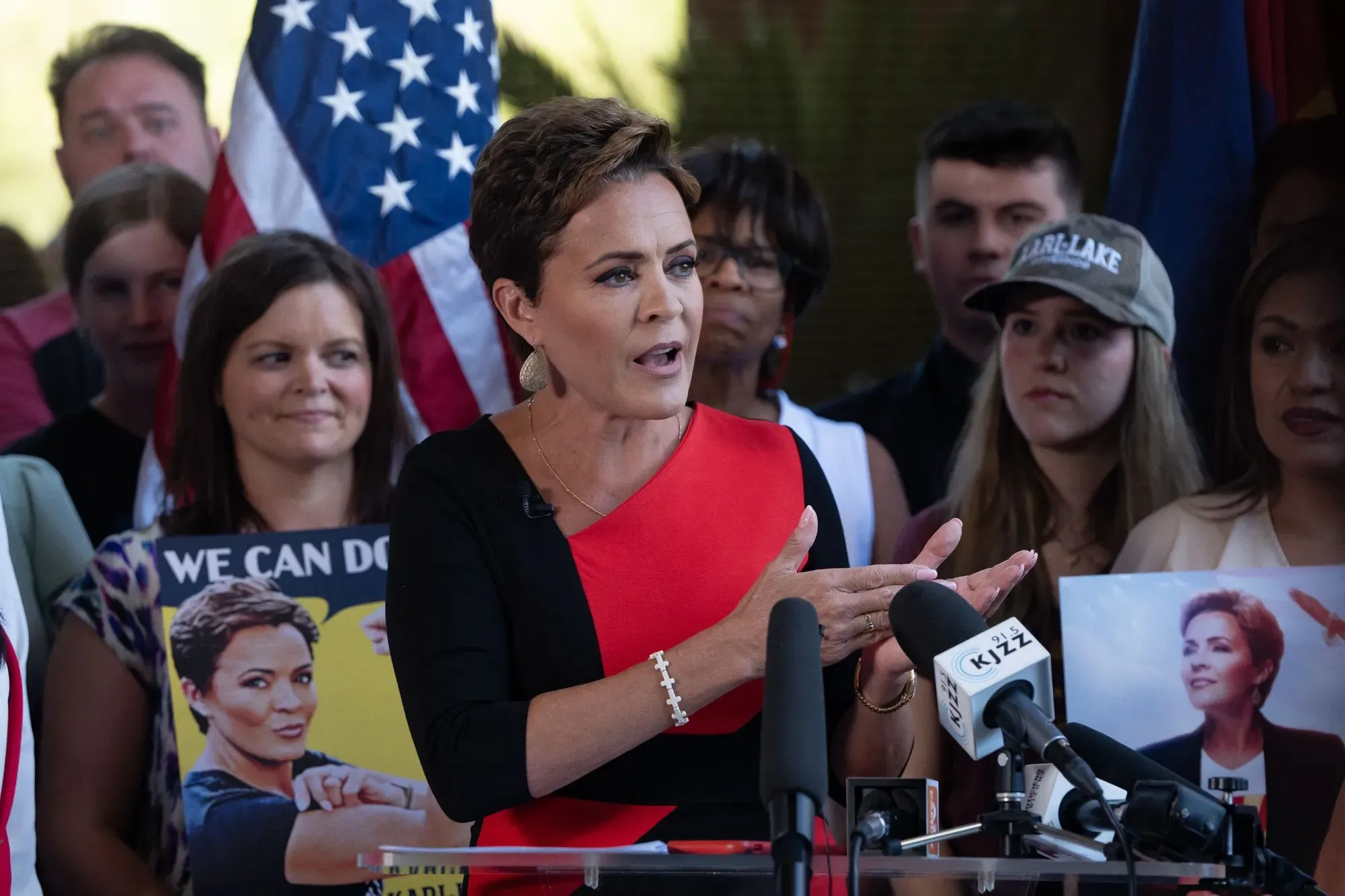 امرأة تتحدث بحماس خلال حدث سياسي، مرتدية فستانًا أحمر، بينما يظهر خلفها مؤيدون يحملون لافتات وصورًا.