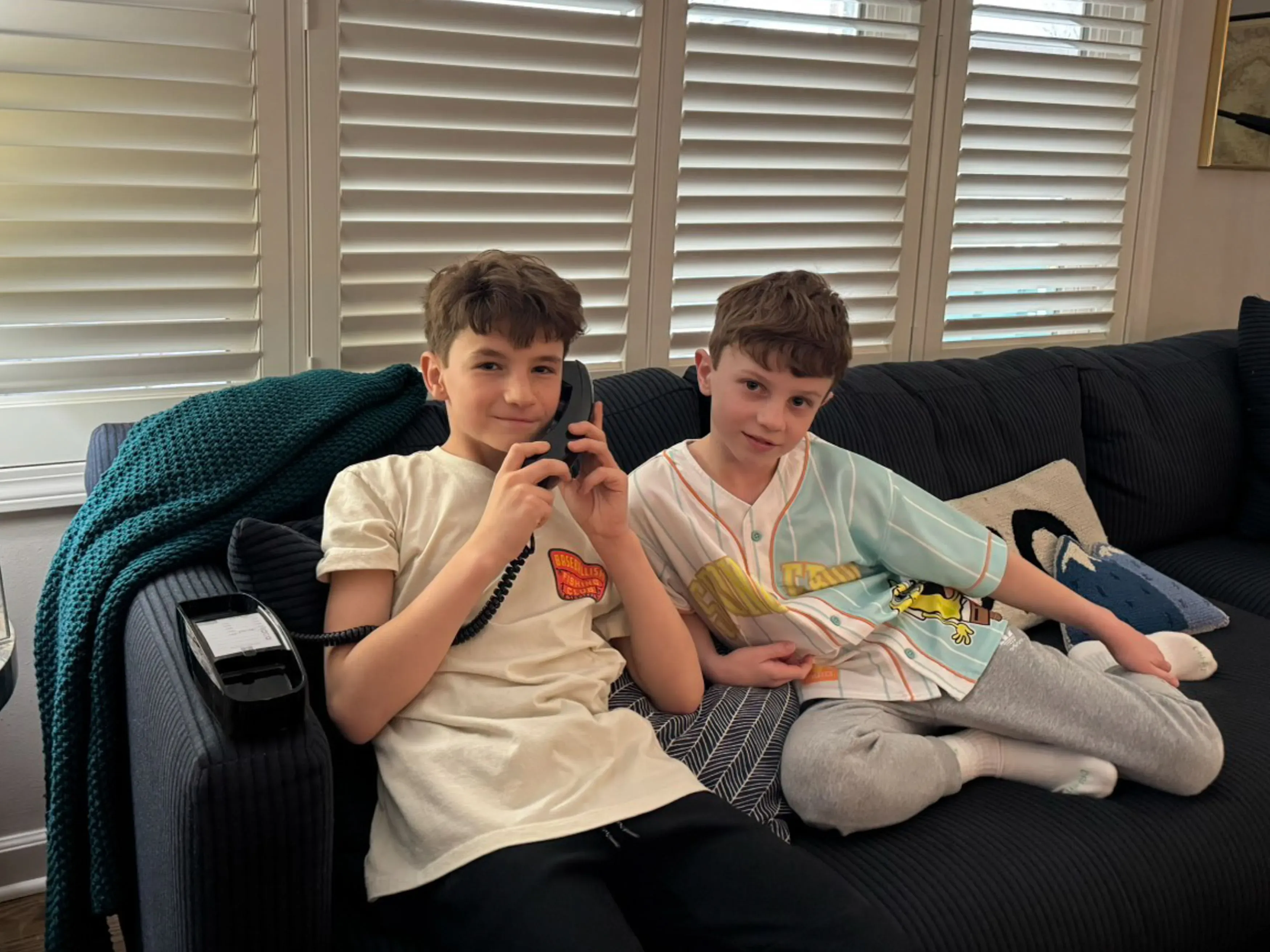 صبيان يجلسان على أريكة، أحدهما يتحدث في هاتف أرضي. تعكس الصورة أهمية التواصل التقليدي للأطفال في عصر التكنولوجيا.