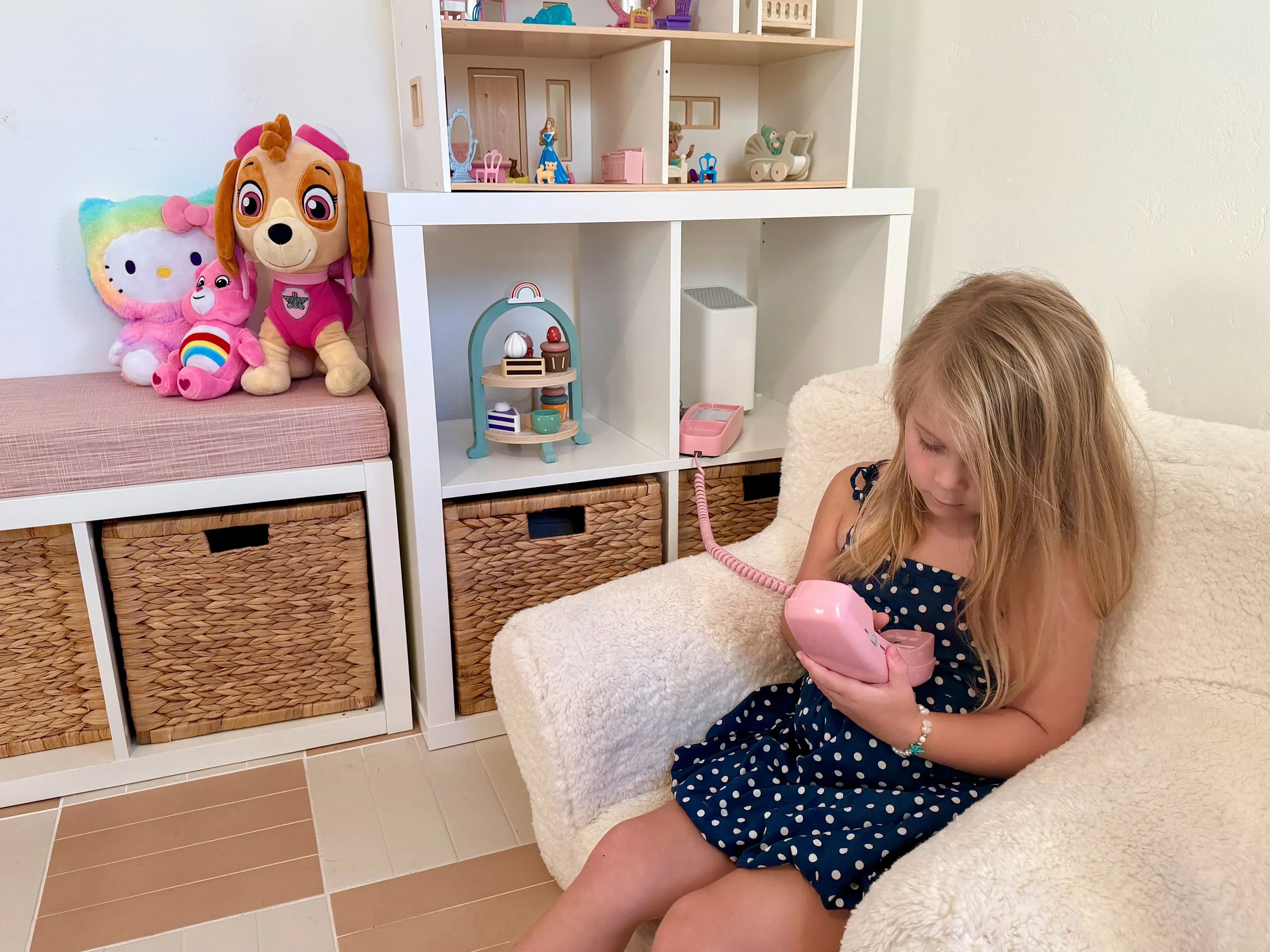 فتاة صغيرة تجلس على أريكة تحمل هاتفًا أرضيًا ورديًا، محاطة بألعابها. تعكس الصورة أهمية التواصل العائلي في عصر التكنولوجيا.