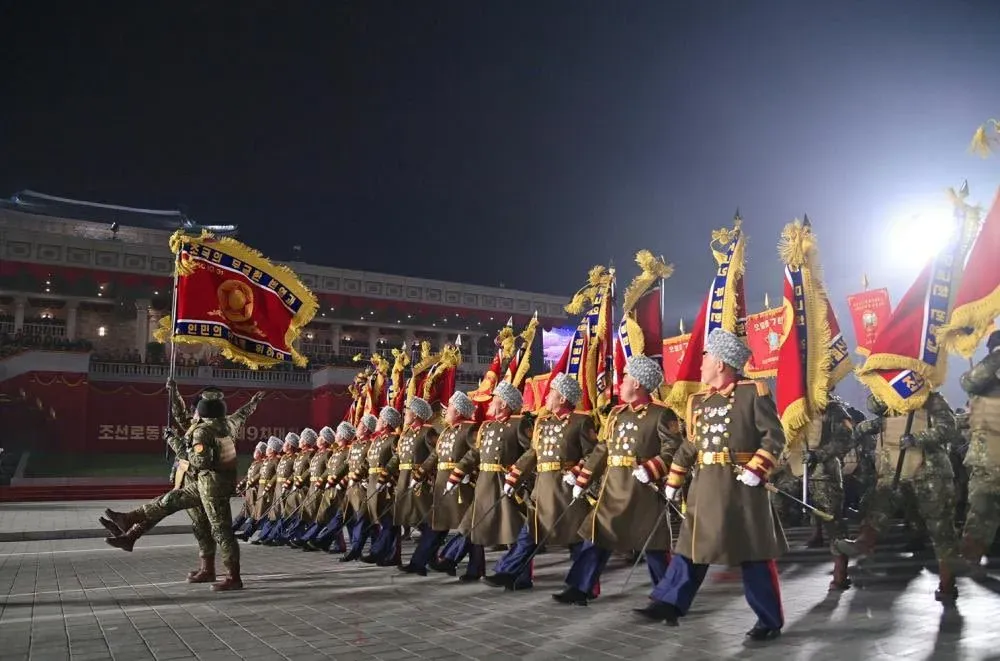 جنود كوريون شماليون يرتدون زيًا رسميًا ويستعرضون الأعلام خلال عرض عسكري ليلي في ساحة كيم إيل سونغ ببيونغ يانغ، مع إضاءة قوية.