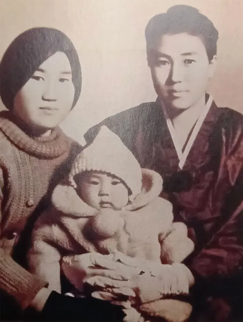 صورة عائلية قديمة تظهر امرأة وطفلًا في ملابس دافئة، مع رجل يرتدي زيًا تقليديًا، تعكس تاريخ التبني في كوريا الجنوبية.