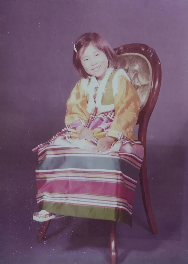 صورة لطفلة كورية ترتدي زيًا تقليديًا وتبتسم، تجلس على كرسي في استوديو تصوير، تعكس ذكريات مؤلمة لعائلات مفصولة.