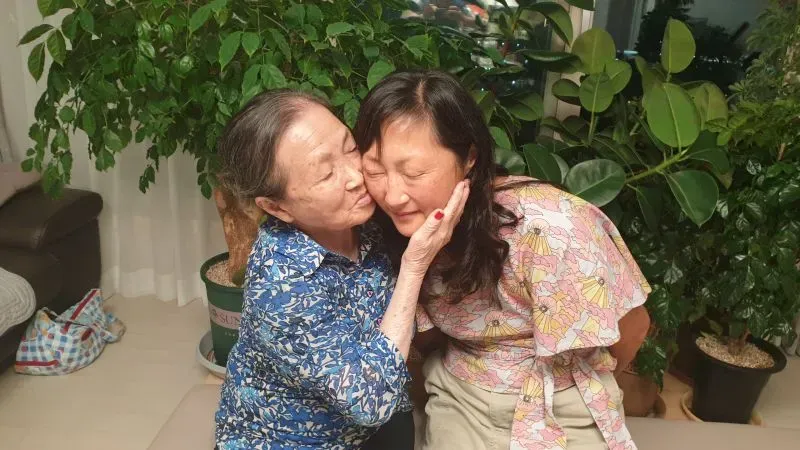 امرأة مسنّة تعانق امرأة شابة في غرفة مليئة بالنباتات، تعبيرات وجههما تحمل مشاعر الحب والحنين.