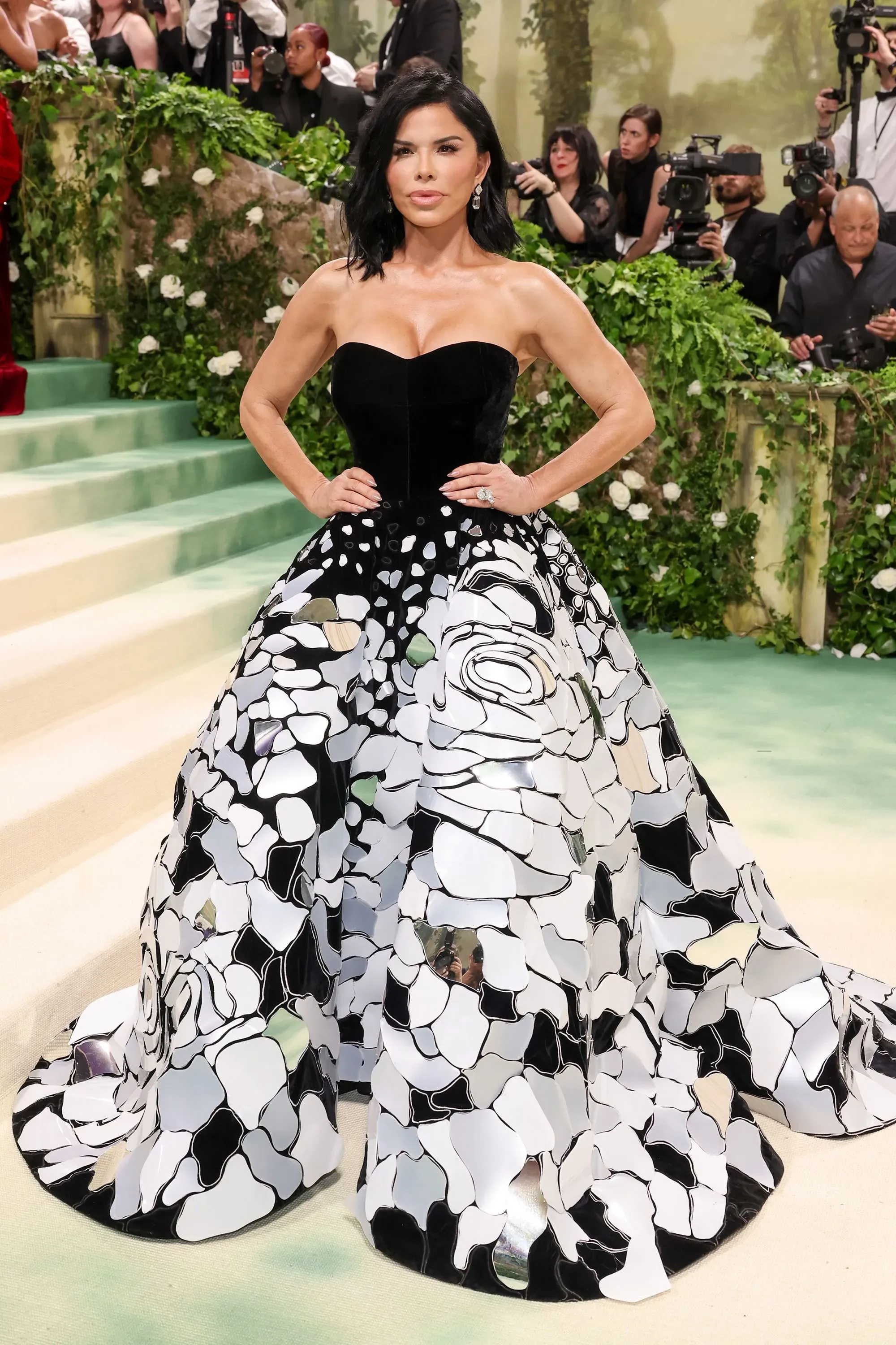 سانشيز بيزوس ترتدي فستاناً أنيقاً بتصميم مميز بالأبيض والأسود خلال حدث الموضة، مع خلفية خضراء وزهور، تعكس اهتمامها بالأزياء الراقية.