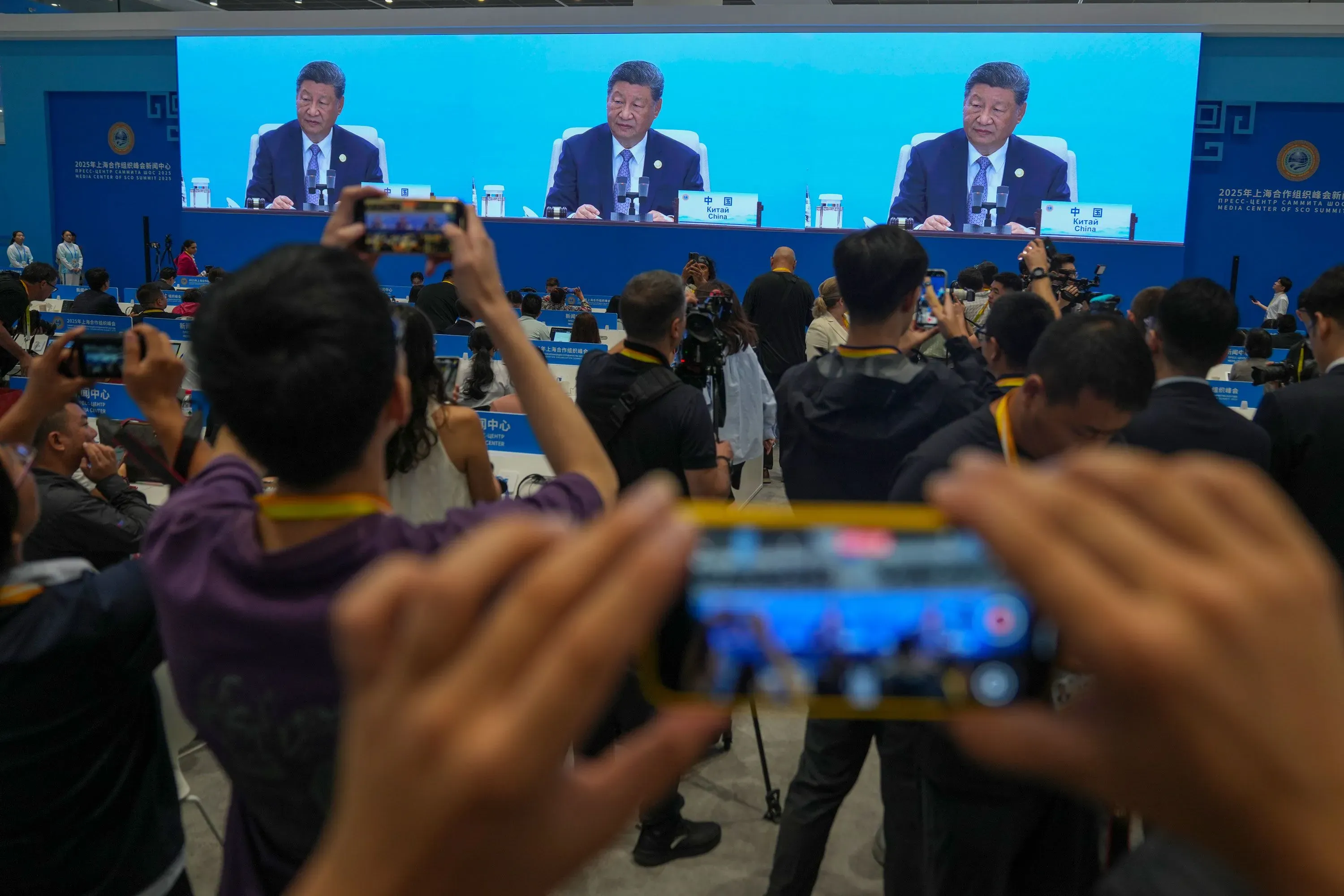 شي جين بينغ يتحدث في قمة منظمة شنغهاي للتعاون، محاطاً بالصحفيين والمشاركين، مع عرض كبير خلفه يعكس أهمية الحدث.