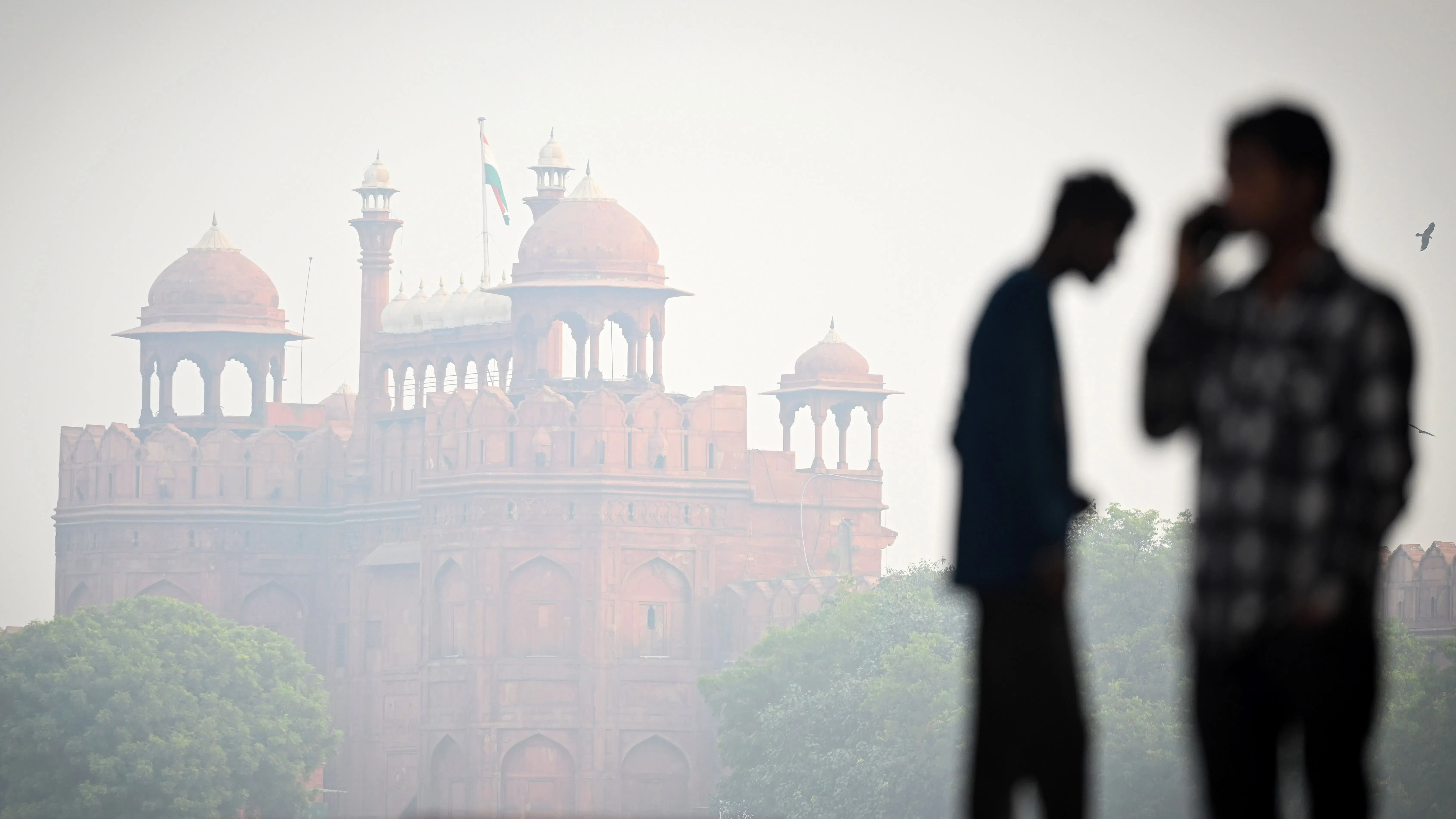 ضباب دخاني كثيف يغلف القلعة الحمراء في نيودلهي، مع silhouettes لشابين يتحدثان، مما يعكس أزمة تلوث الهواء الخطيرة.