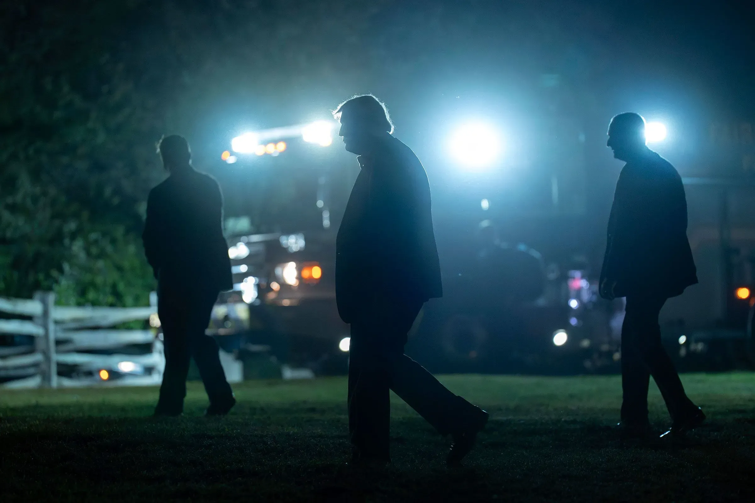 ظلال ثلاثة رجال يسيرون في الليل، مع إضاءة خلفية قوية من مصابيح سيارات، تعكس أجواء مشحونة في سياق المناخ السياسي المتوتر.