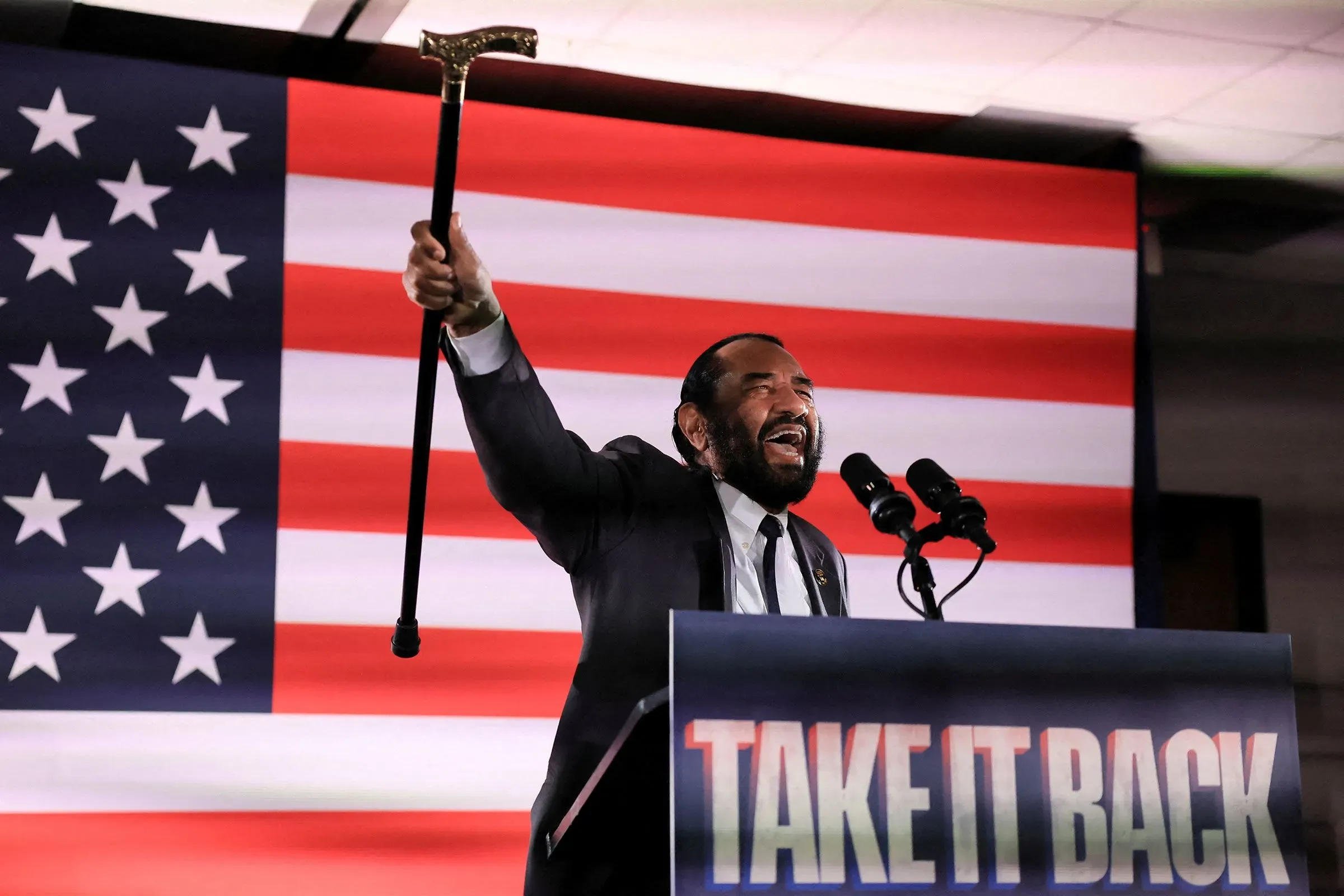 رجل يرفع عصا ويعبر عن حماسه خلال خطاب انتخابي أمام علم أمريكي، في سياق الانتخابات في الدائرة الثامنة عشرة بولاية تكساس.