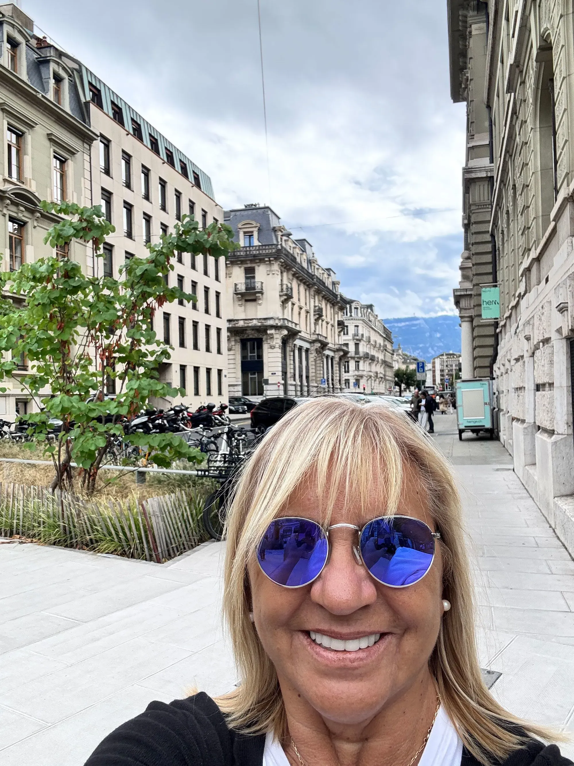 امرأة تبتسم في صورة سيلفي، ترتدي نظارات شمسية زرقاء، وتقف في شارع يضم مباني تاريخية وأشجار، مما يعكس تأثير الذكاء الاصطناعي على المترجمين.