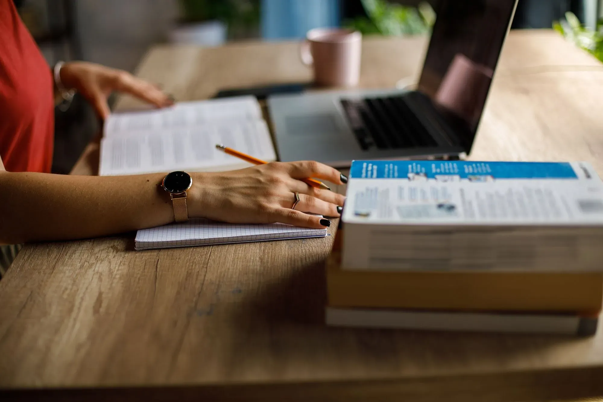 امرأة تكتب في دفتر ملاحظات بجانب كمبيوتر محمول، مع كتب مرجعية على الطاولة، تعكس تأثير الذكاء الاصطناعي على مهنة الترجمة.