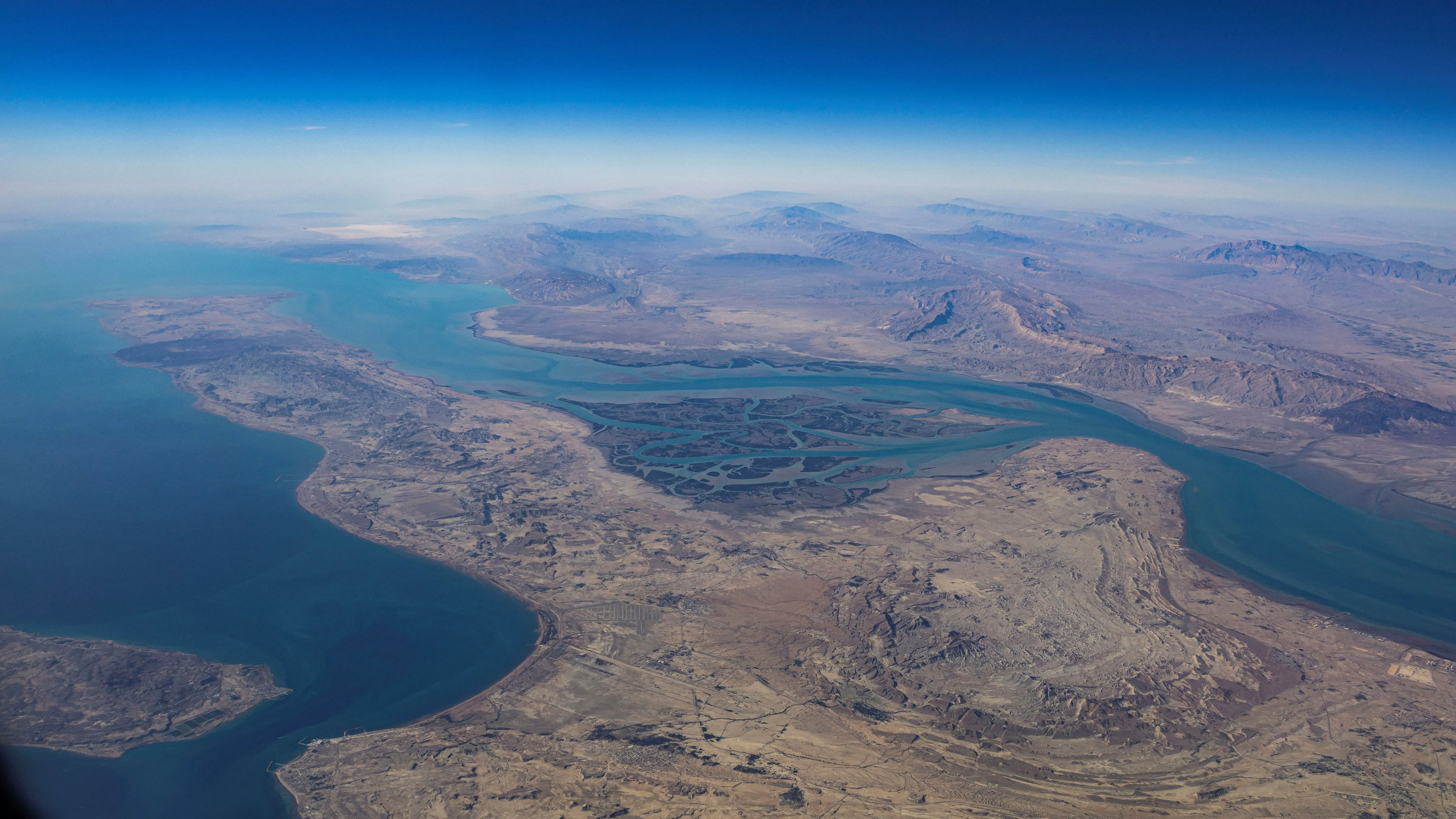 منظر جوي لمضيق هرمز، يظهر المياه الزرقاء العميقة والساحل الجبلي الإيراني، يمثل نقطة اختناق حيوية لنقل النفط والغاز عالميًا.