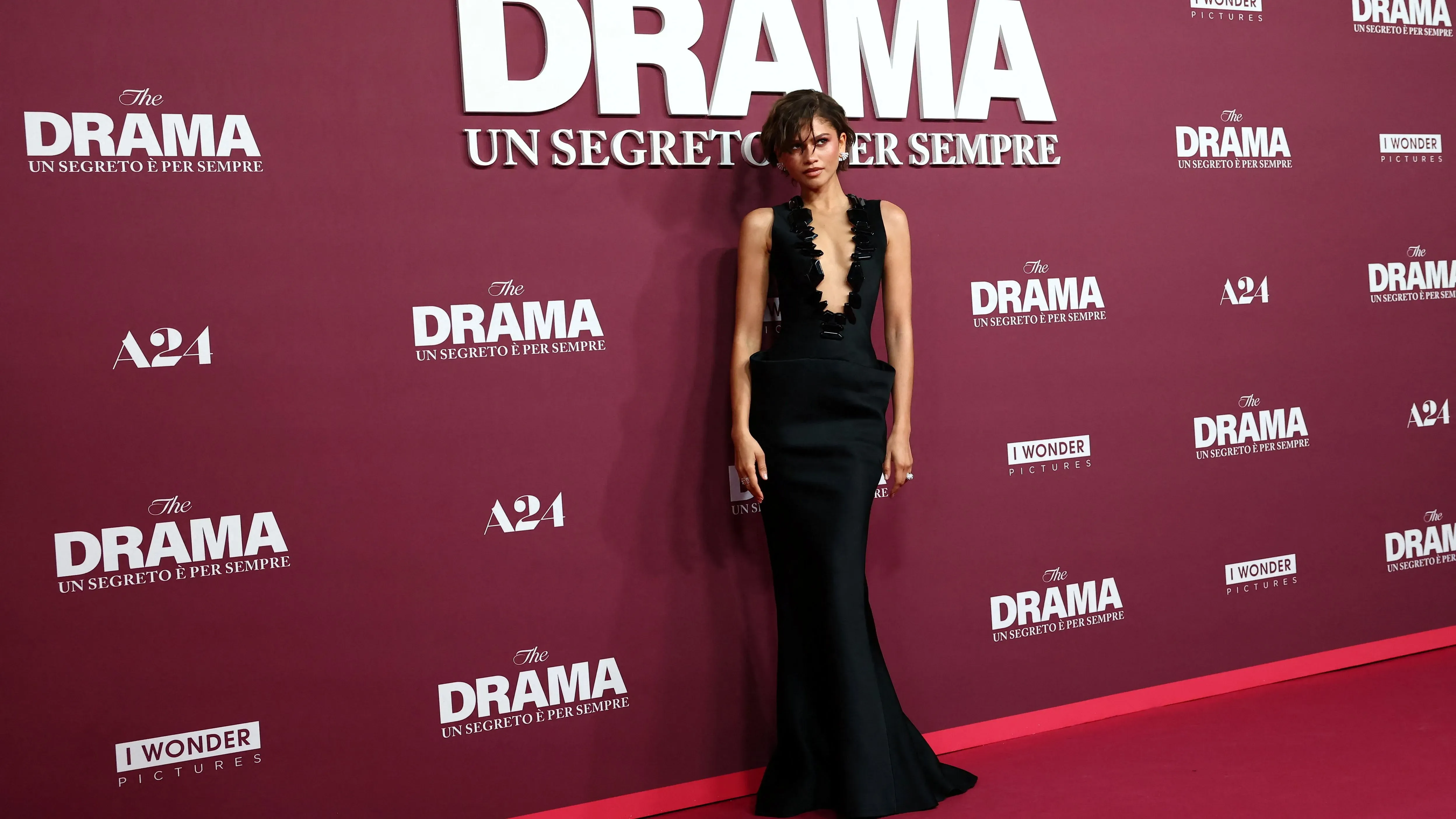 زندايا ترتدي فستانًا أسود ضيقًا من أرماني في العرض الأول لفيلم "ذا دراما"، مع خلفية تحمل اسم الفيلم وشعار A24.