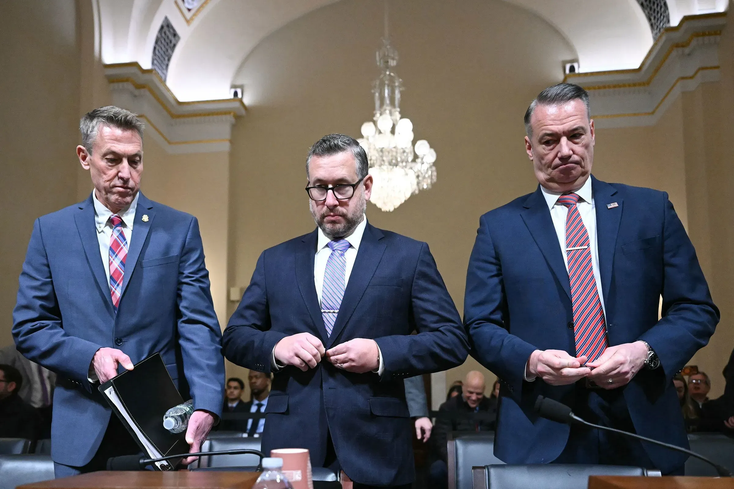 ثلاثة مسؤولين حكوميين يرتدون بدلات رسمية، يقفون في قاعة جلسة استماع، مع تعبيرات وجه تعكس القلق والجدية، في سياق تحقيقات تتعلق بإبشتاين.