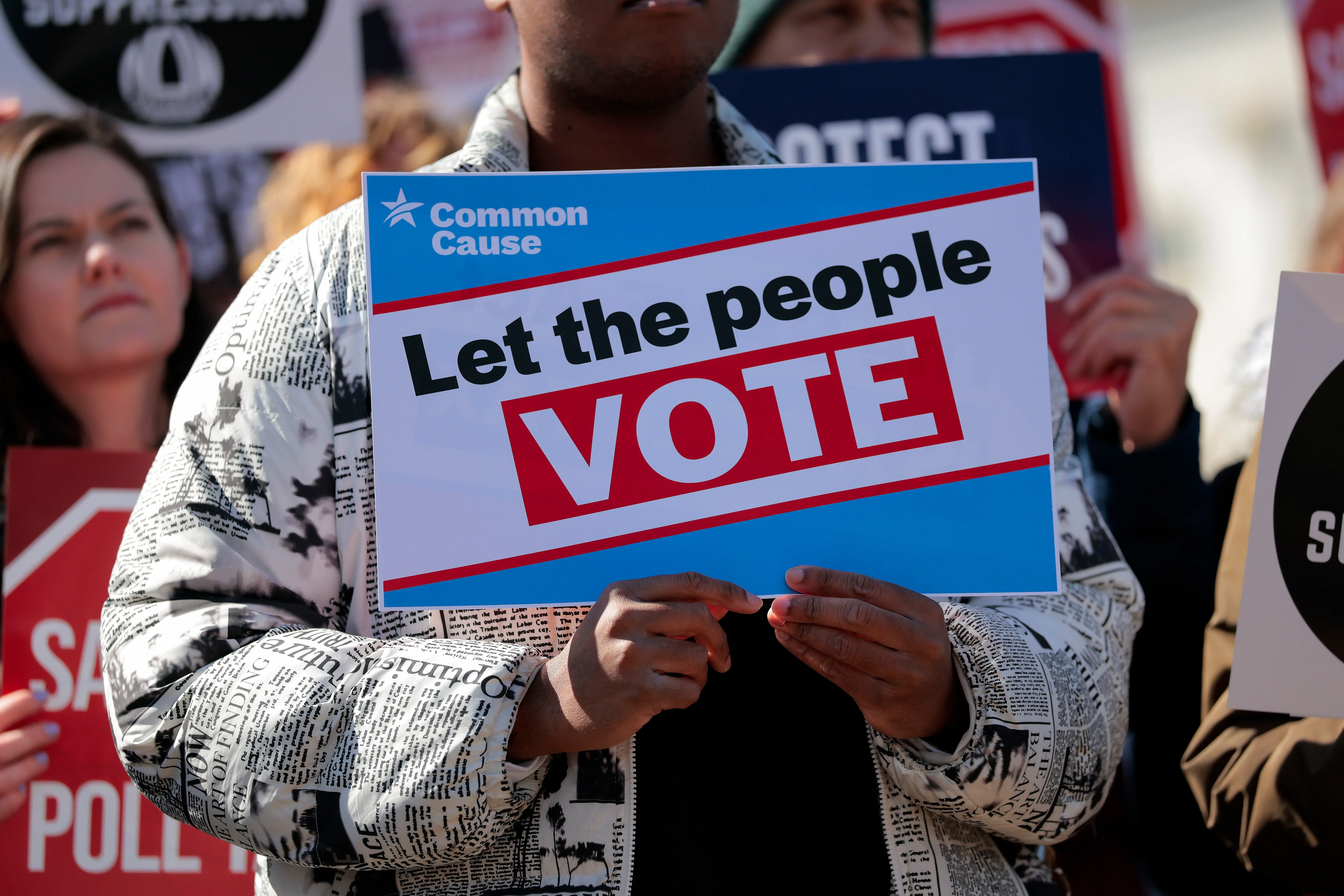 لافتة في تجمع احتجاجي تحمل عبارة "دعوا الناس يصوتون"، مع متظاهرين في الخلفية يدعمون حقوق التصويت.