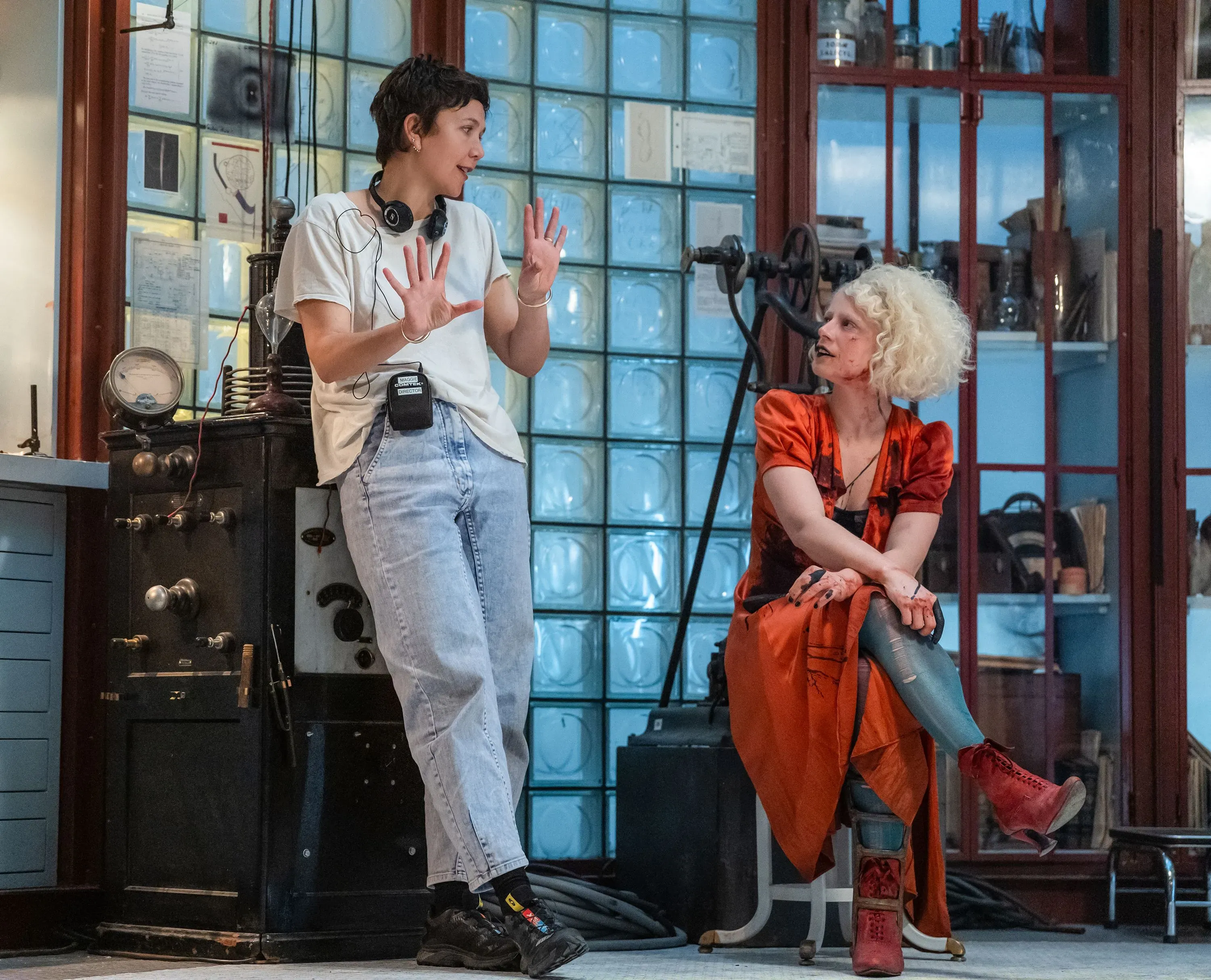 مخرجة الفيلم "العروس!" ماغي جيلينهال تتحدث مع الممثلة جيسي باكلي في موقع التصوير، مع خلفية تحتوي على معدات سينمائية.