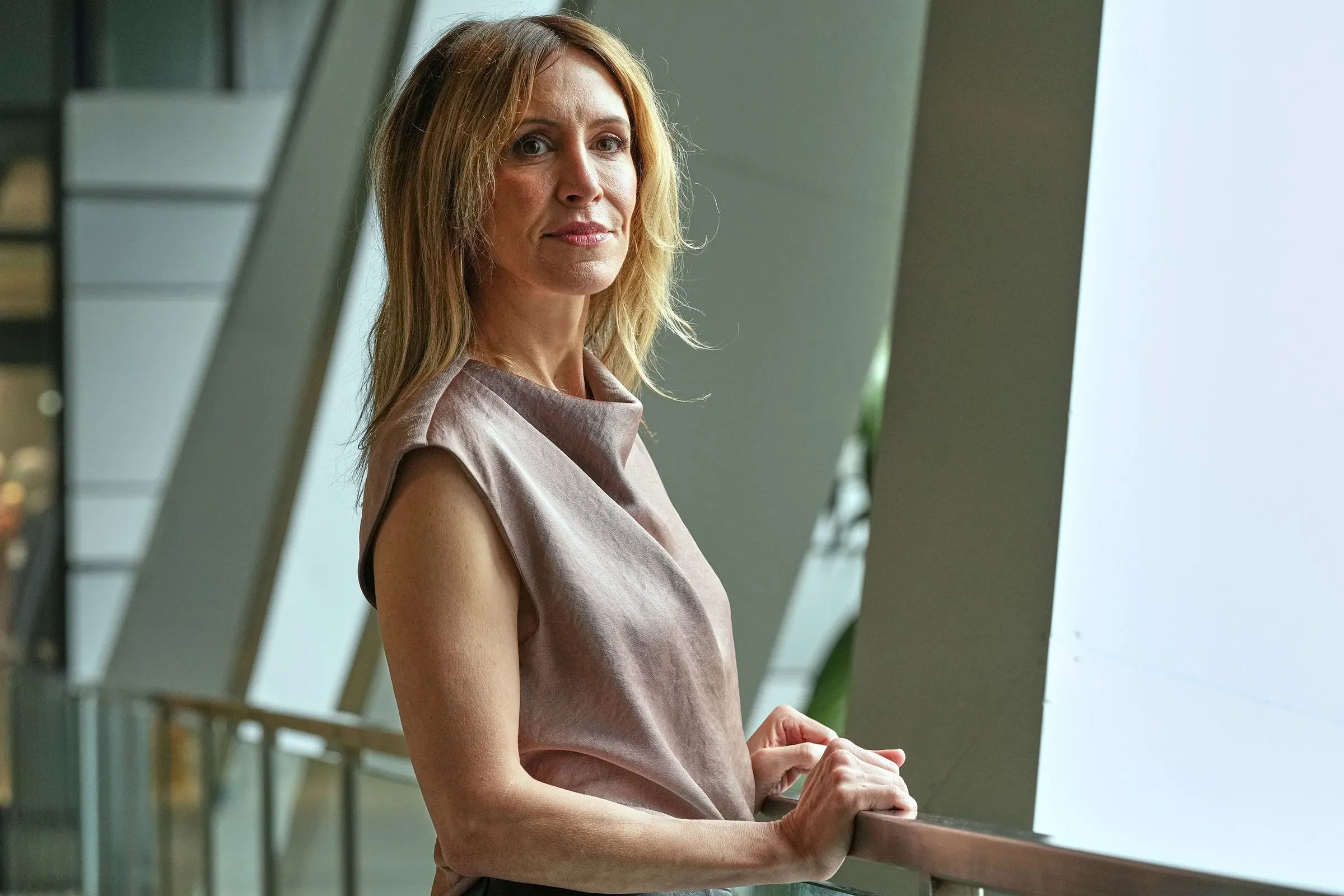 امرأة تقف بجانب درابزين في مكان داخلي حديث، تعبر عن الثقة والتصميم، مما يعكس اهتمامها بقضايا الصحة والبيئة.