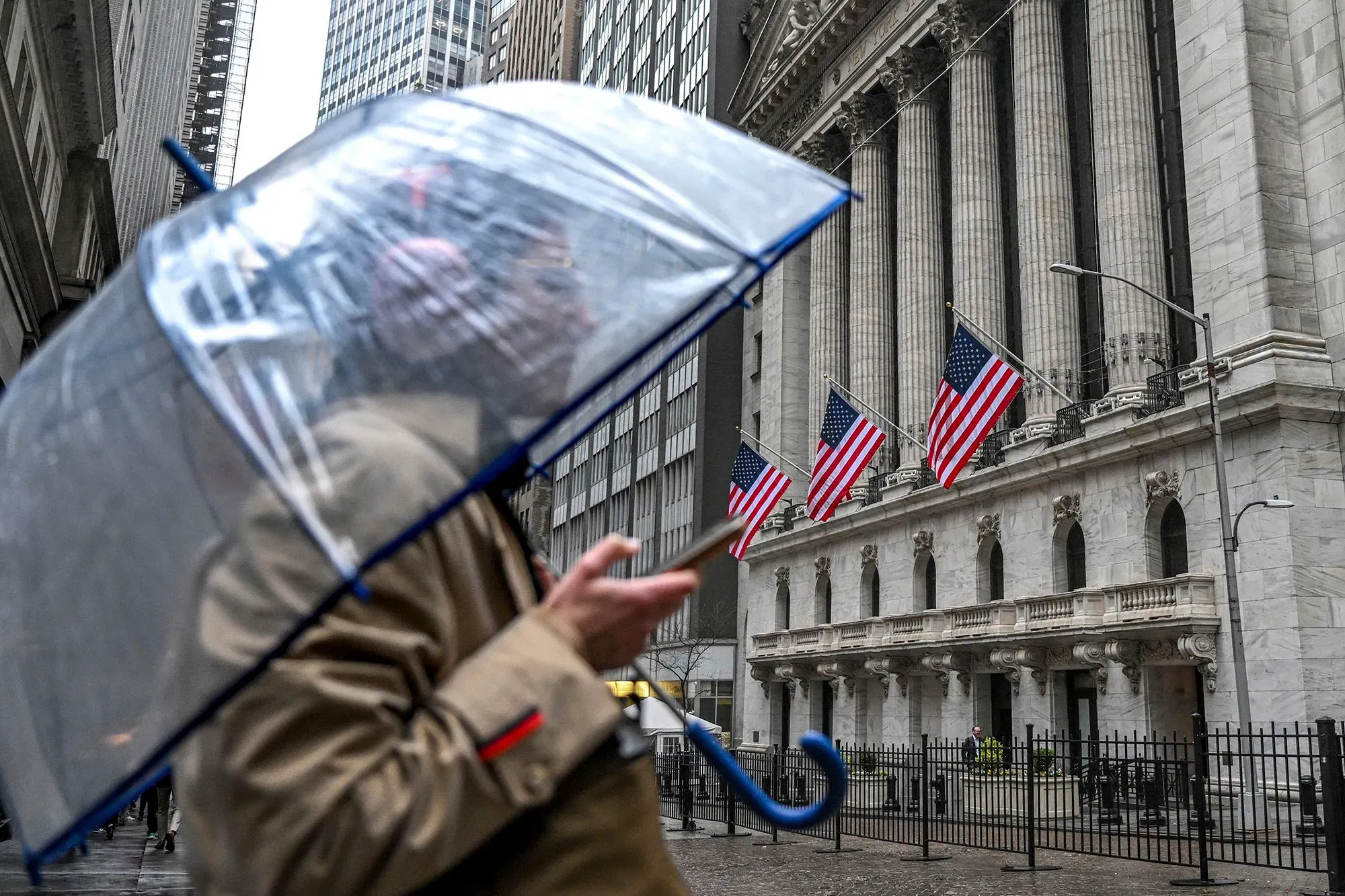 شخص يحمل مظلة شفافة أمام مبنى البورصة الأمريكية، مع أعلام الولايات المتحدة ترفرف في الخلفية، مما يعكس أجواء السوق المتقلبة.