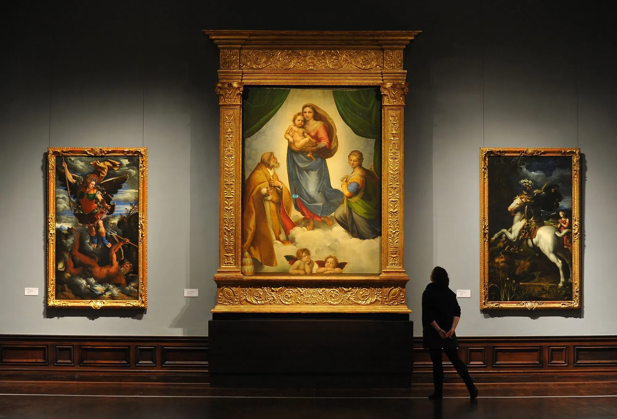 لوحة فنية كبيرة في متحف، تُظهر مشهدًا دينيًا مع شخصيات ملونة، بينما يقف زائر يتأملها.