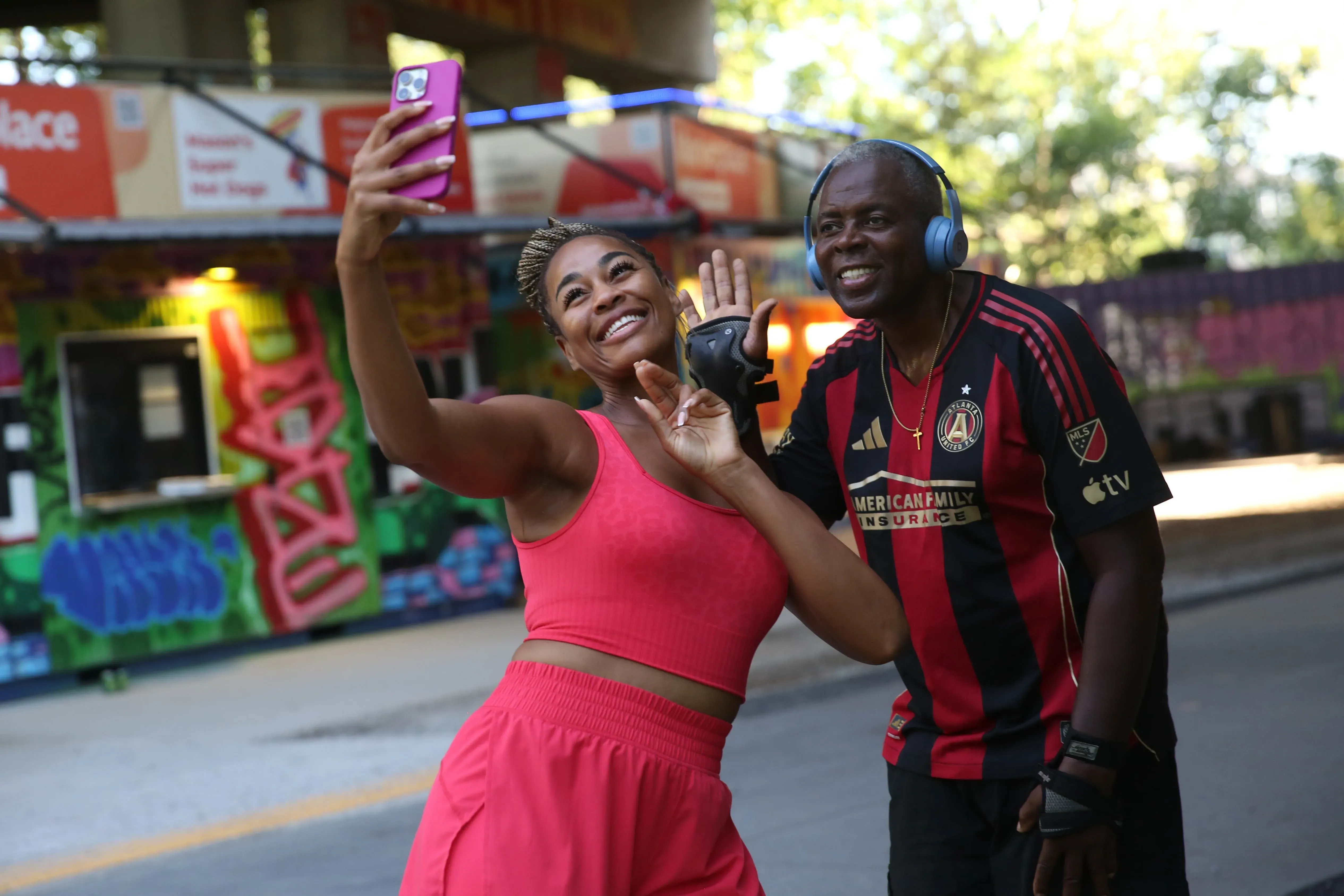 امرأة ترتدي ملابس رياضية وردية تلتقط سيلفي مع رجل يرتدي قميص فريق أتلانتا. خلفهما جدران مزينة برسومات جرافيتي، مما يعكس حيوية بيلت لاين.