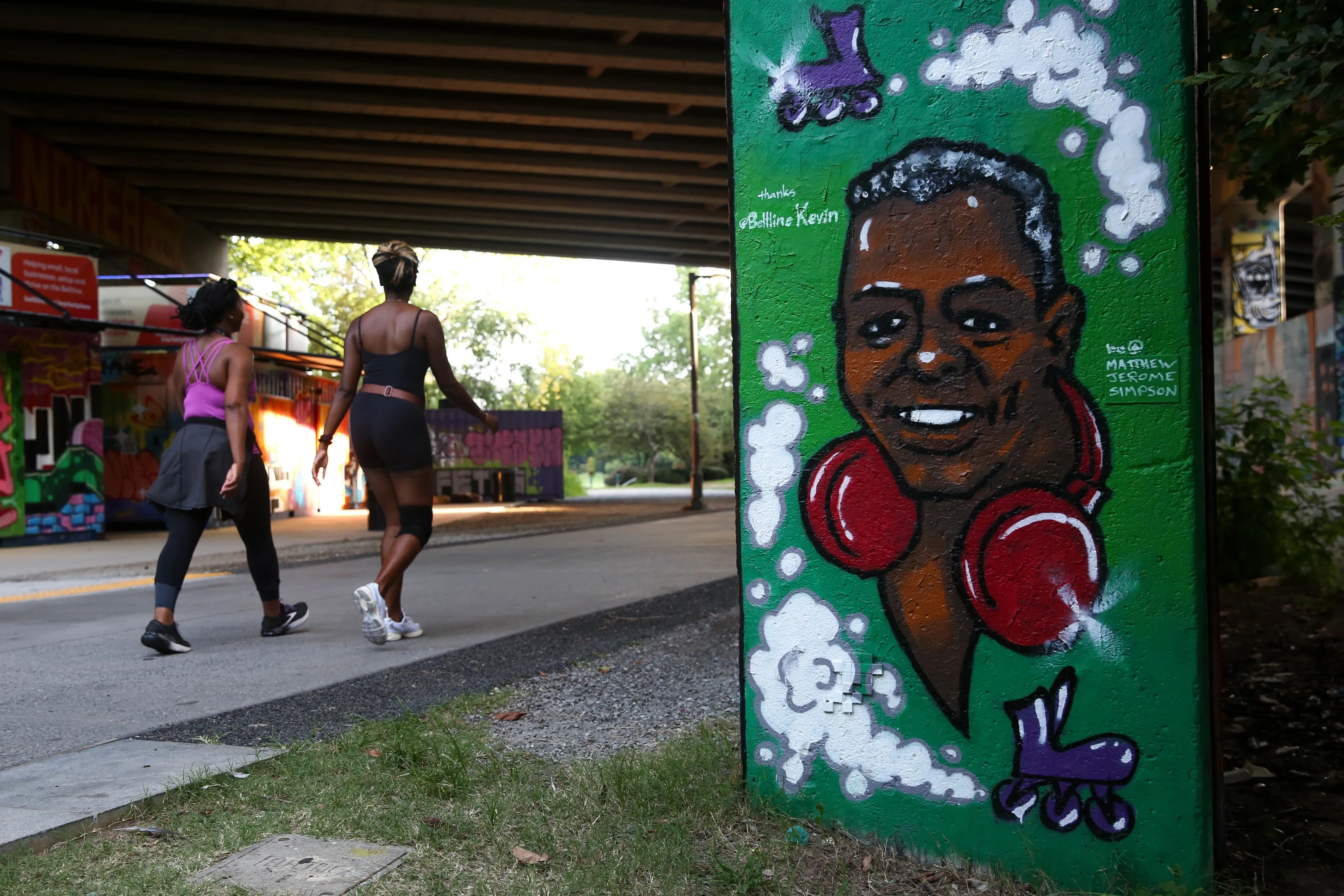 رسم جدارية ملونة تظهر وجه "كيفن بيلت لاين" مبتسمًا مع سماعات رأس حمراء، بينما تسير امرأتان بجانبه في ممر بيلت لاين بأتلانتا.