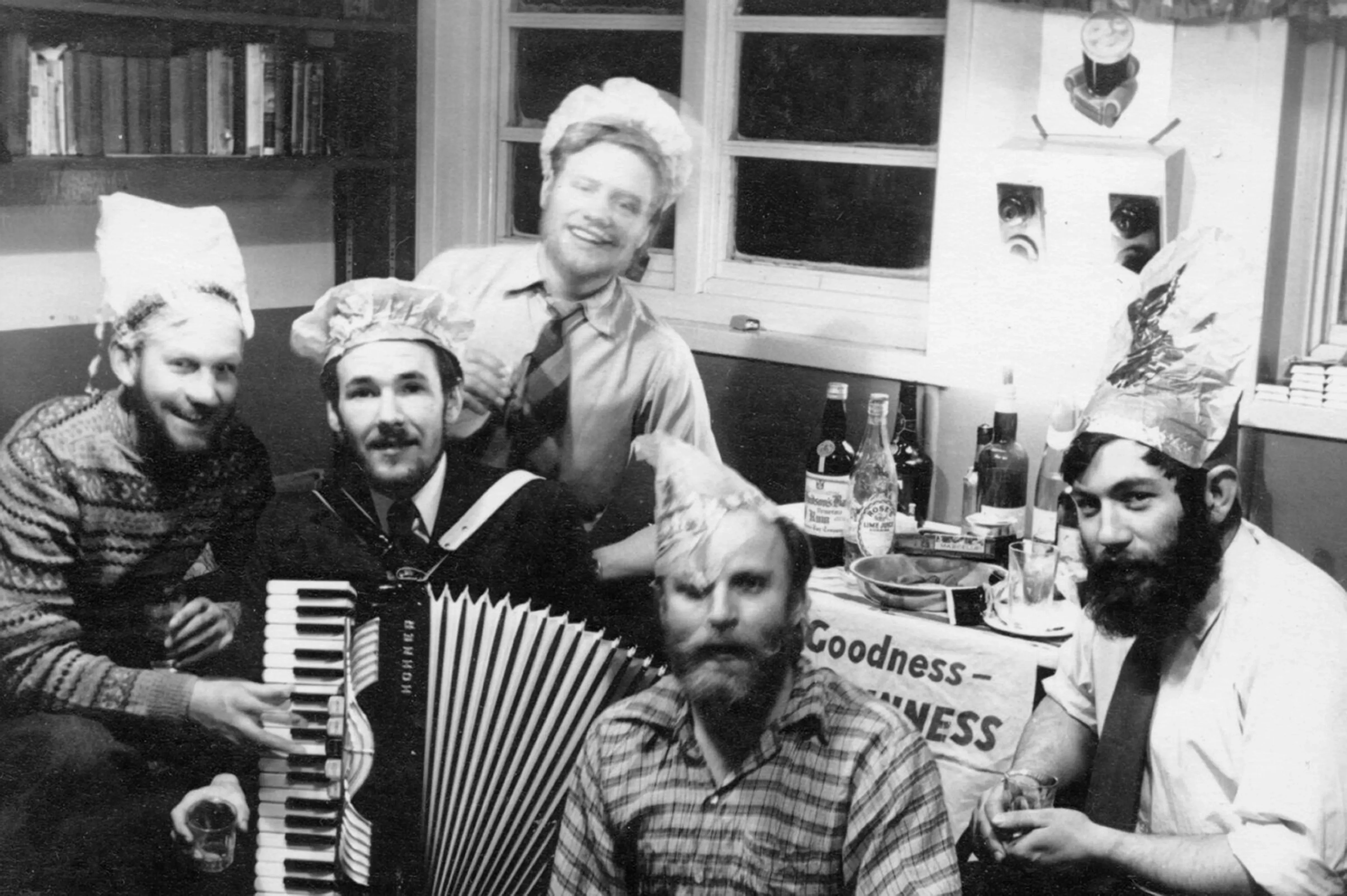 مجموعة من الرجال يجلسون معًا في غرفة، يرتدون قبعات ورقية، ويحتفلون، مع أكورديون وزجاجات مشروبات خلفهم.