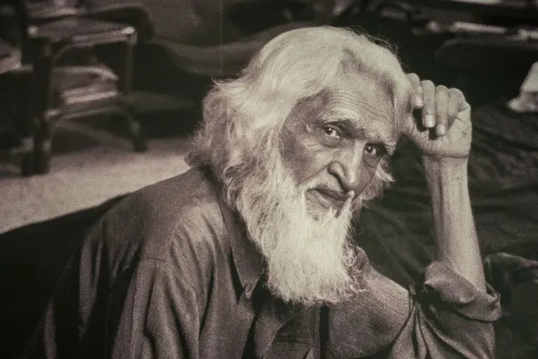 صورة فنية تظهر مقبول فدا حسين، الفنان الهندي الشهير، ذو اللحية البيضاء، وهو يجلس بتعبير عميق في بيئة داخلية.