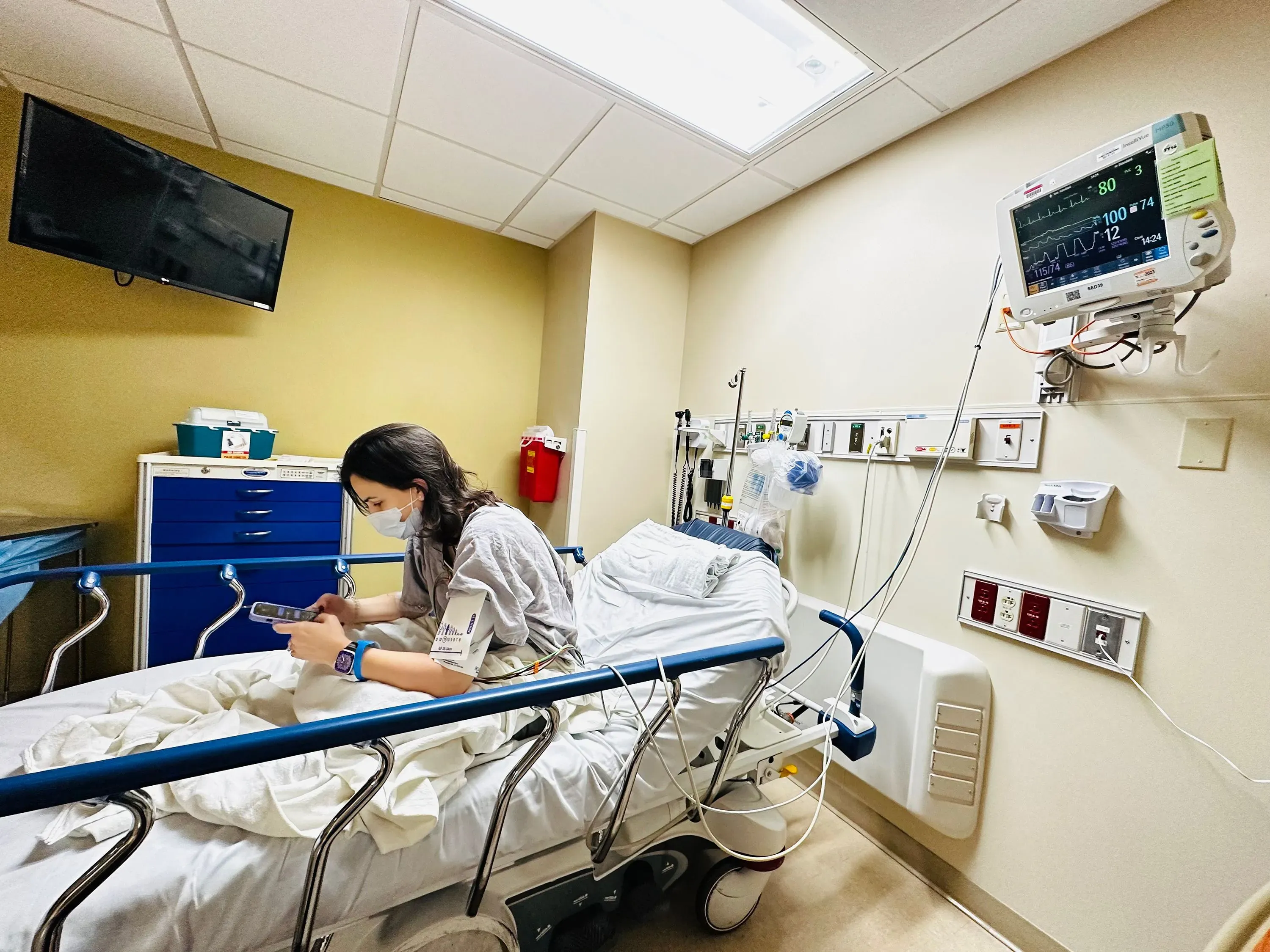 امرأة شابة تجلس على سرير في غرفة مستشفى، ترتدي كمامة وتستخدم هاتفها، مع وجود أجهزة طبية تراقب حالتها.