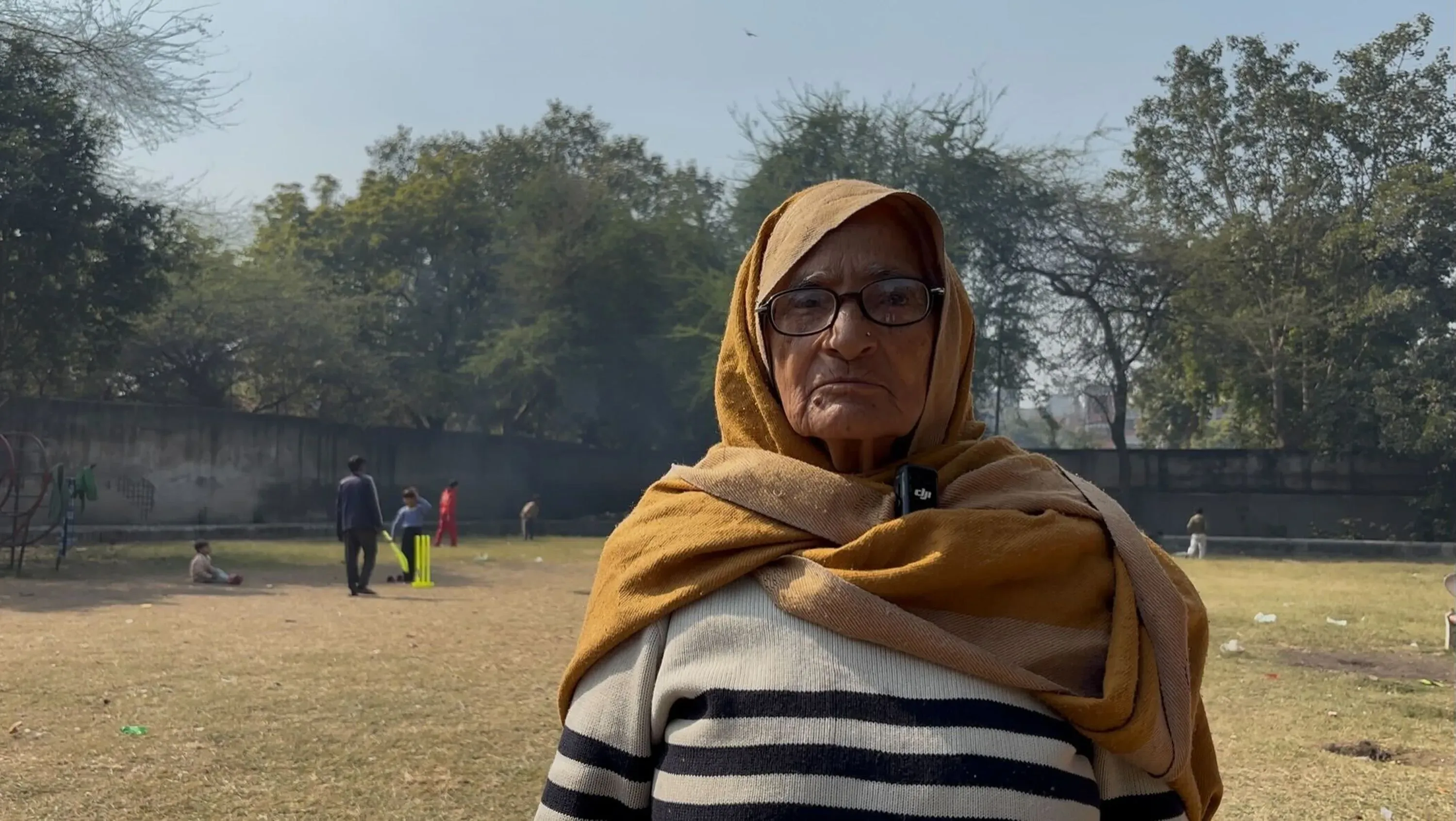 امرأة مسنّة ترتدي شالاً وتظهر في حديقة، محاطة بأشخاص يلعبون. تعكس الصورة تأثير أزمة المياه في دلهي على المجتمع.