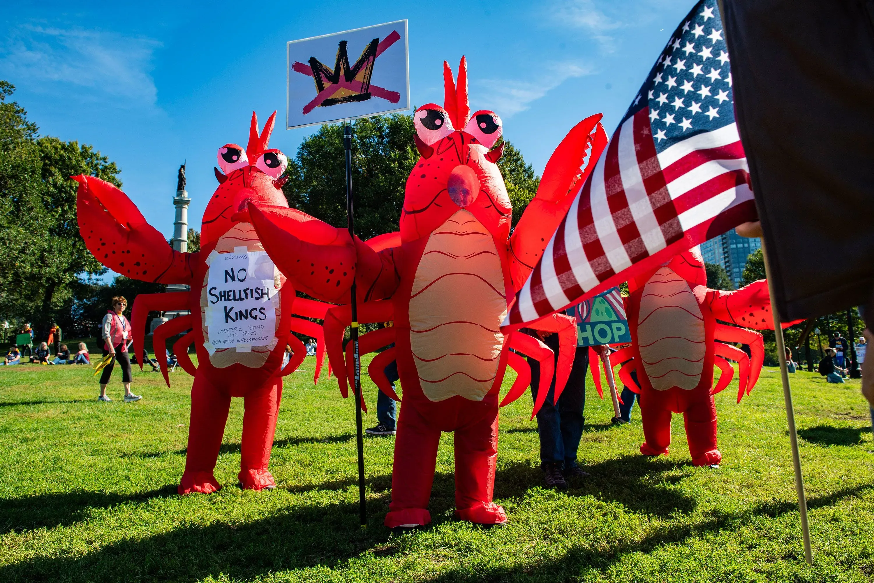 متظاهرون يرتدون أزياء جراد البحر في حديقة، يحملون لافتة "لا للملوك الأنانيين"، مع العلم الأمريكي في الخلفية، خلال احتجاجات ضد إدارة ترامب.