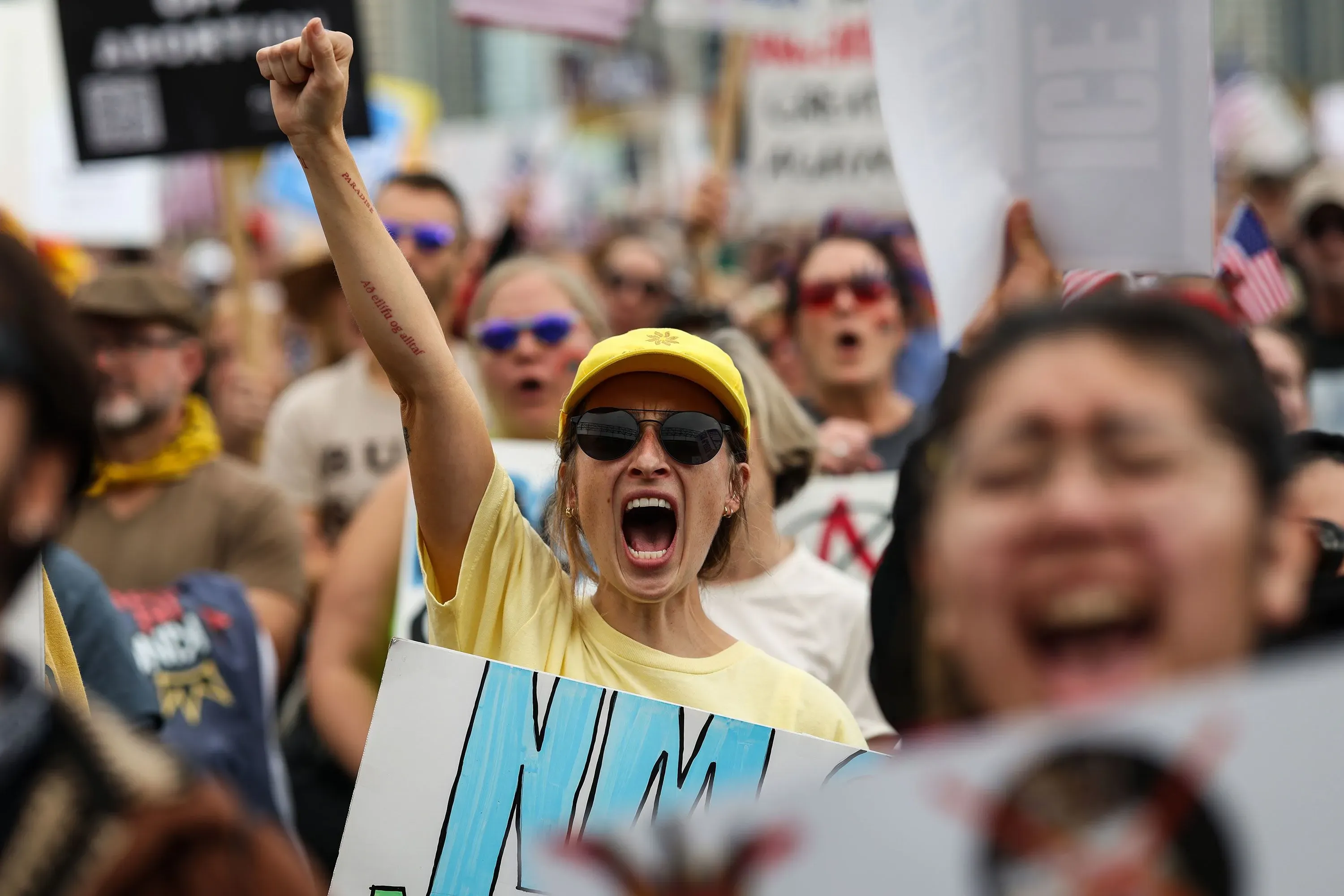 امرأة ترتدي قبعة صفراء ونظارات شمسية، ترفع قبضتها في الهواء أثناء احتجاج حاشد، محاطة بمحتجين آخرين يحملون لافتات تعبر عن معارضتهم لإدارة ترامب.