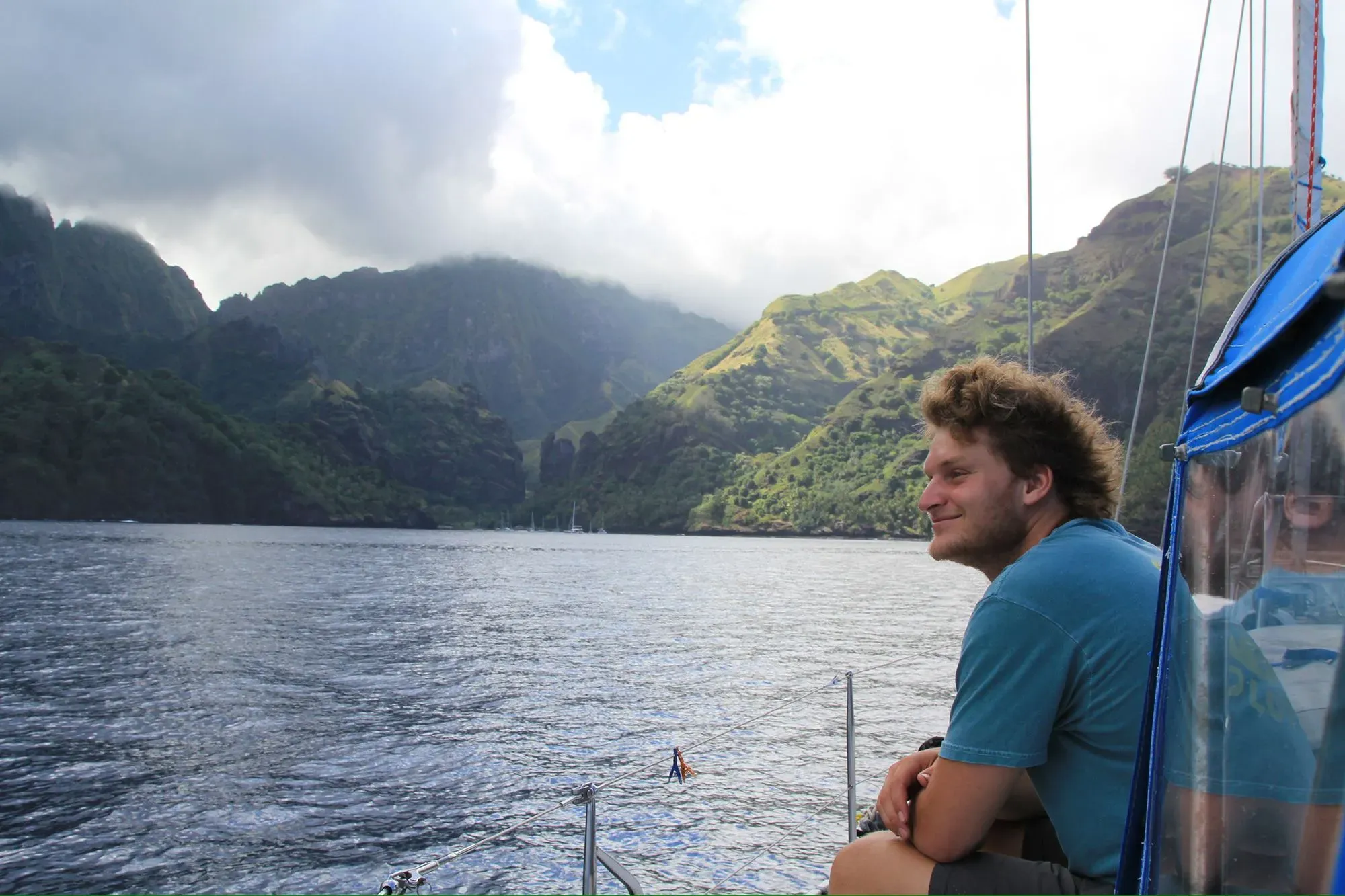 شاب يجلس على حافة قارب شراعي في المحيط، محاطًا بالجبال الخضراء، يعكس روح المغامرة والسعي للحرية.